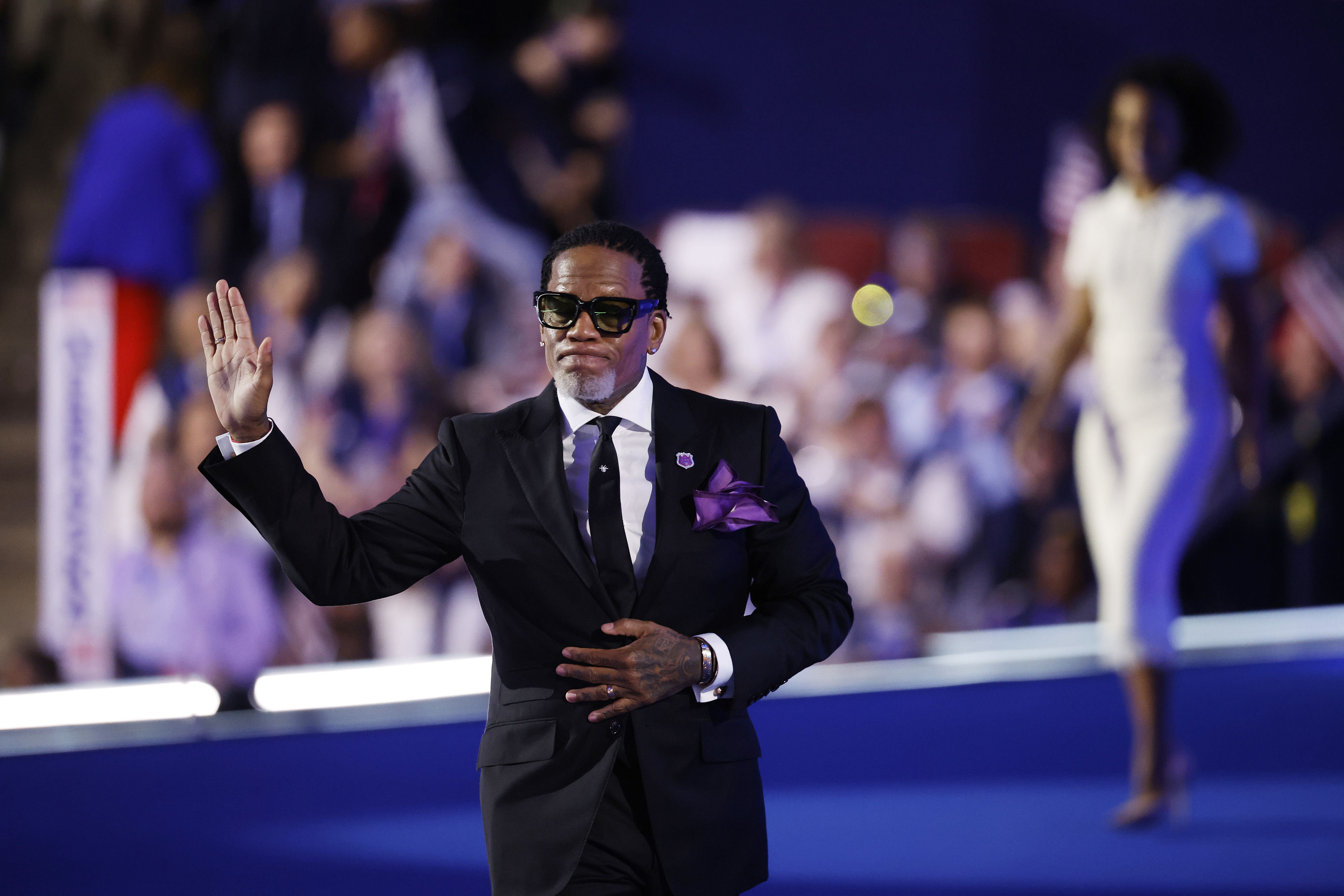 A man in a dark suit and sunglasses waves on a blue stage with a crowd in the background, while a woman in a white dress walks behind him