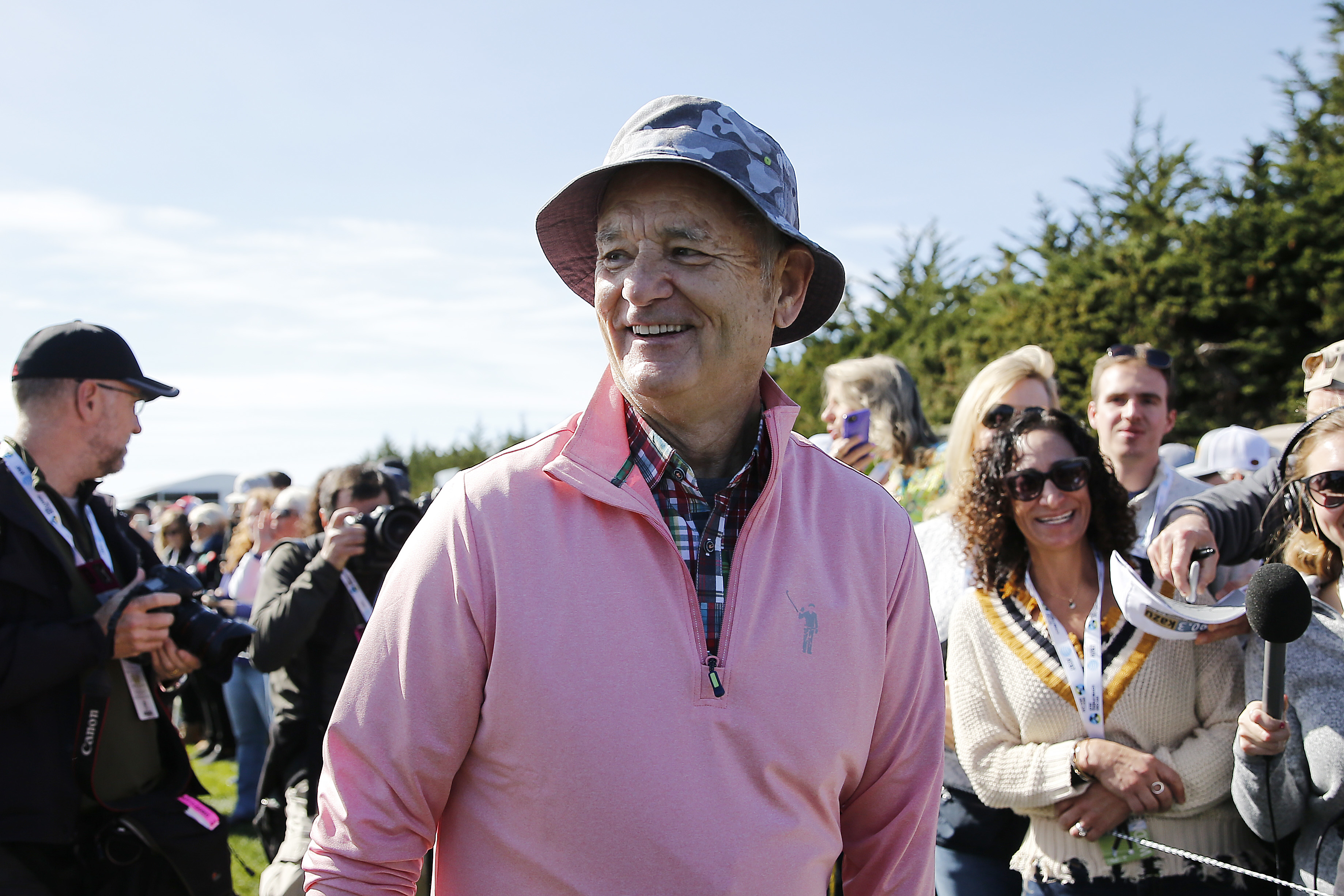 Bill Murray smiles while wearing a hat and casual outdoor clothes, surrounded by fans and photographers at a public event