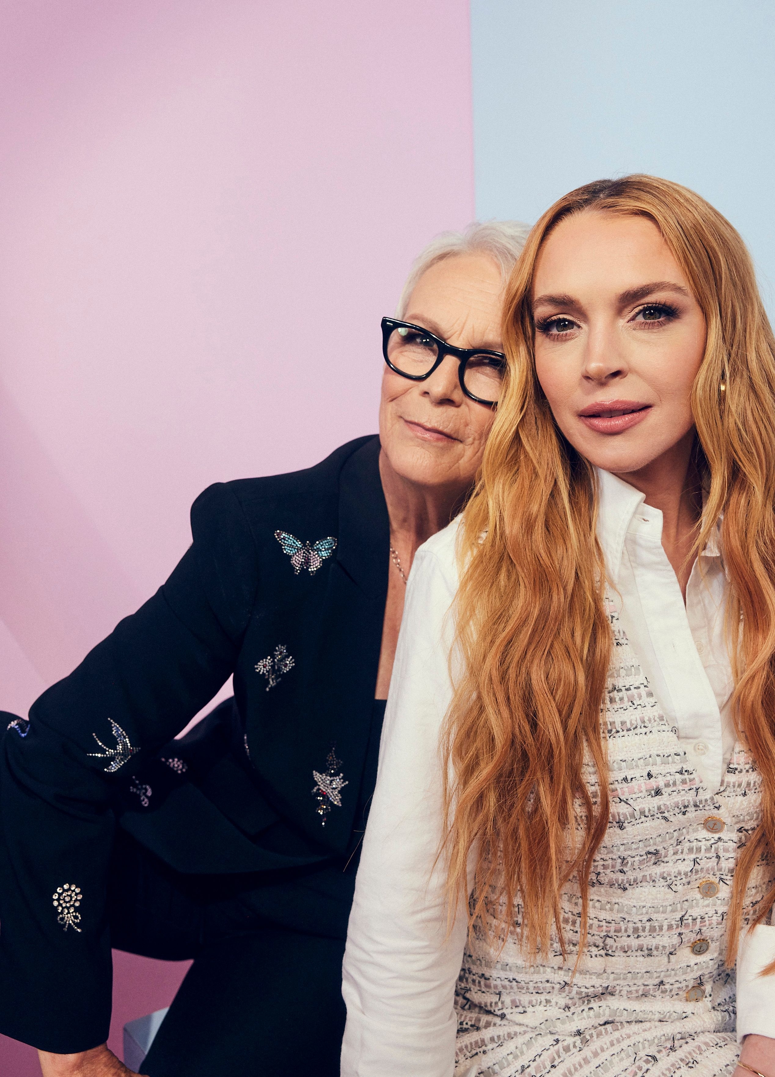 Jamie Lee Curtis, in a butterfly-patterned blazer, stands behind Lindsay Lohan, who wears a white textured dress, both posing for a photo