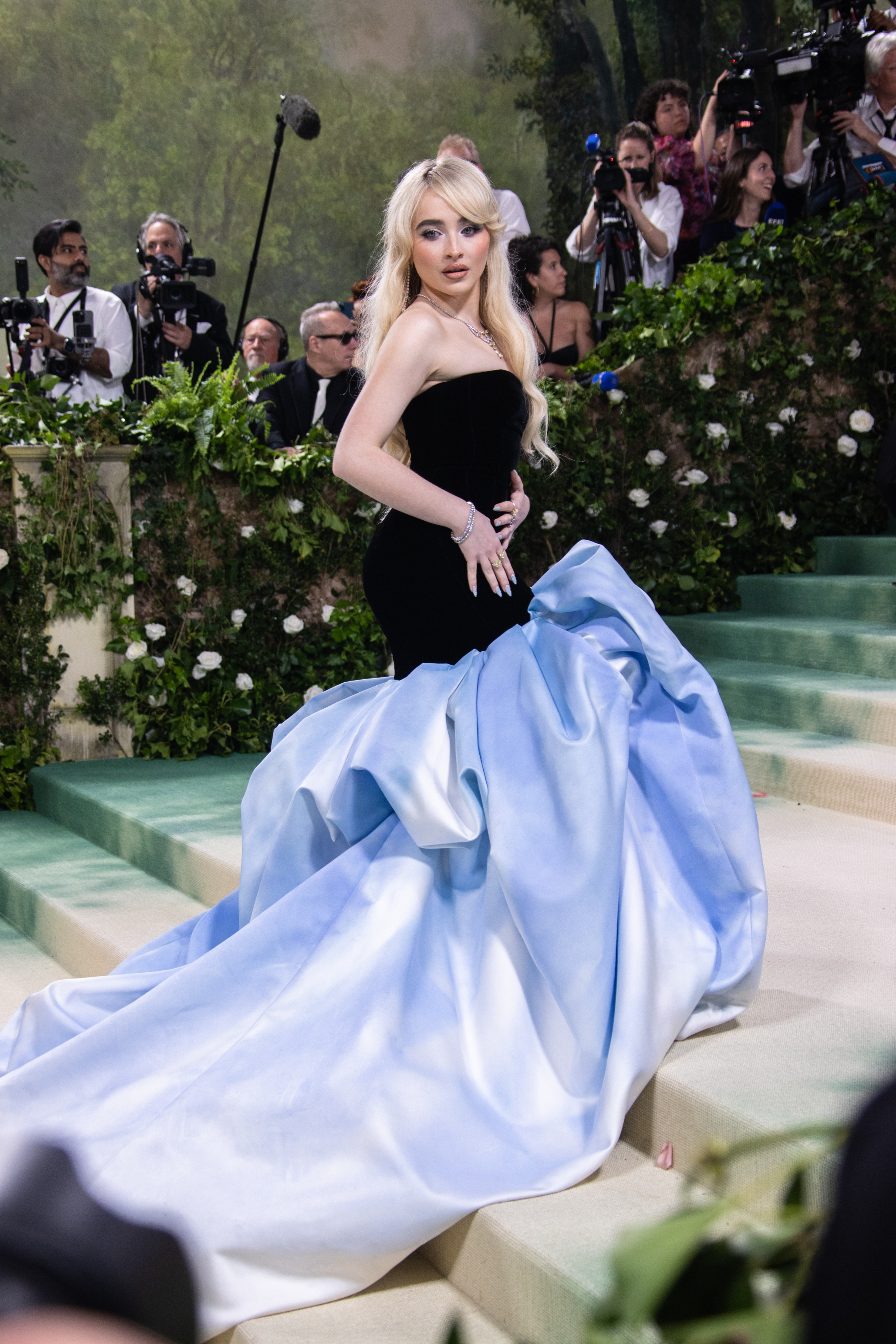 A woman with long blonde hair wearing a sophisticated black top with a dramatic light blue gown poses on steps surrounded by photographers and greenery