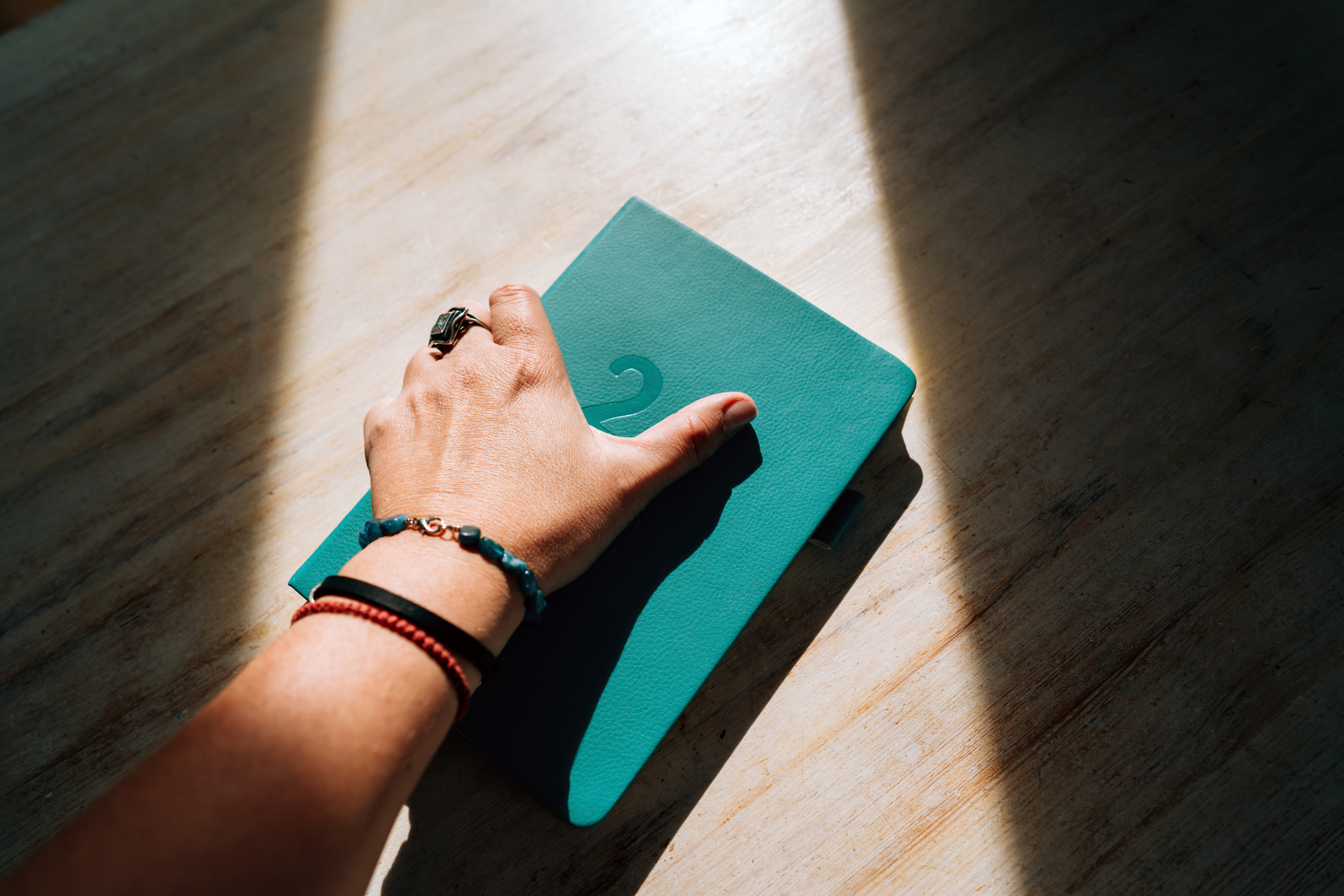 A hand with bracelets and ring is holding a teal notebook on a wooden surface