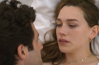 Penn Badgley and Victoria Pedretti are lying close together in bed, gazing into each other's eyes