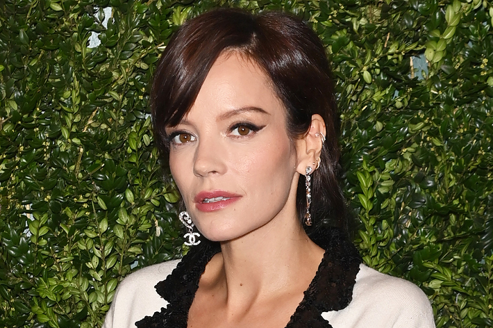 Lily Allen poses against a foliage backdrop, wearing a deep V-neck blazer with black and white trim, accessorized with statement earrings