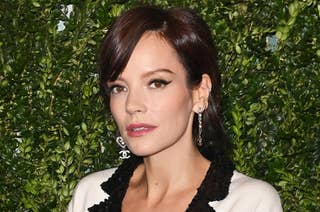 Lily Allen poses against a foliage backdrop, wearing a deep V-neck blazer with black and white trim, accessorized with statement earrings