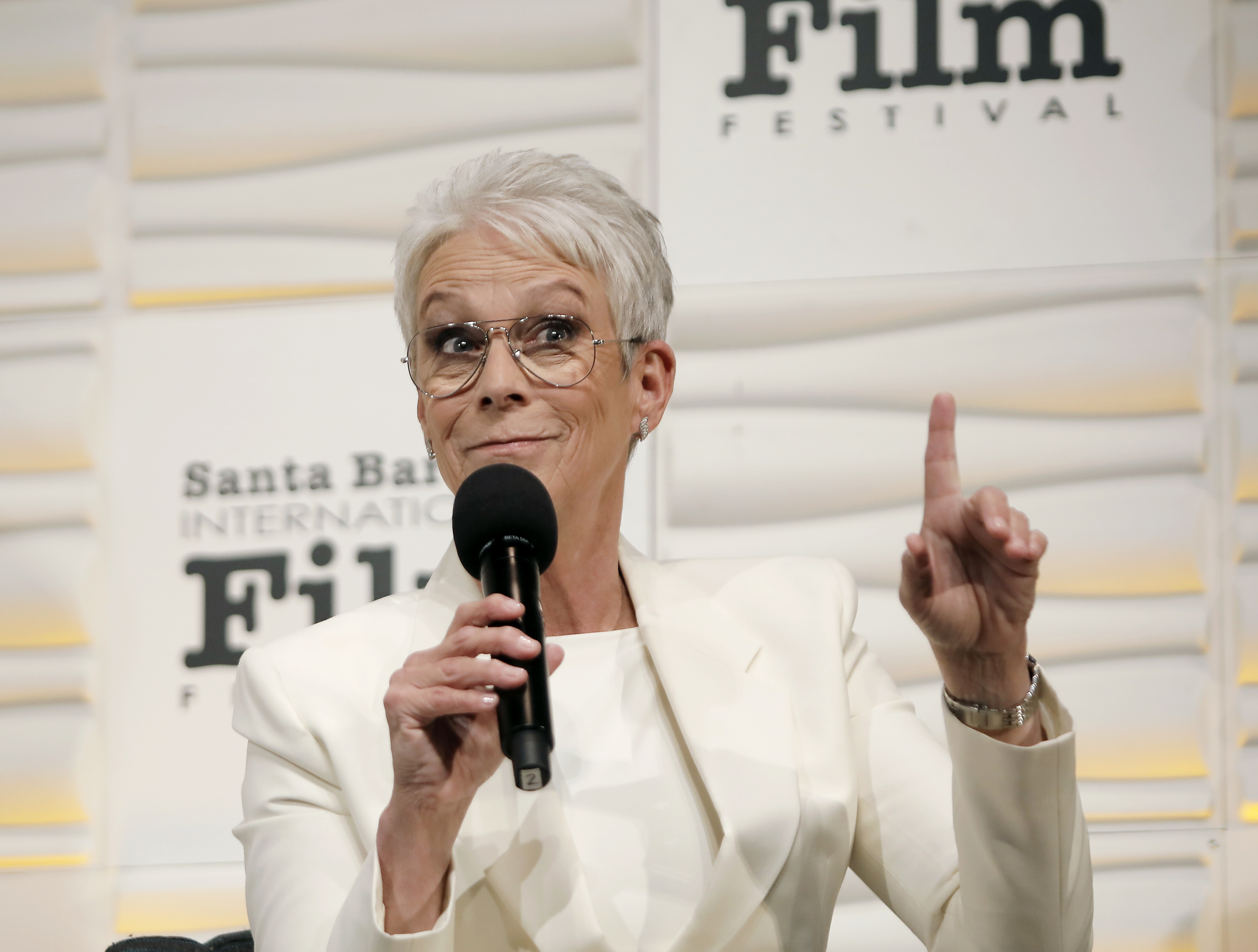 Jamie Lee Curtis speaks into a microphone while raising her finger at the Santa Barbara International Film Festival. She is wearing a white suit and glasses