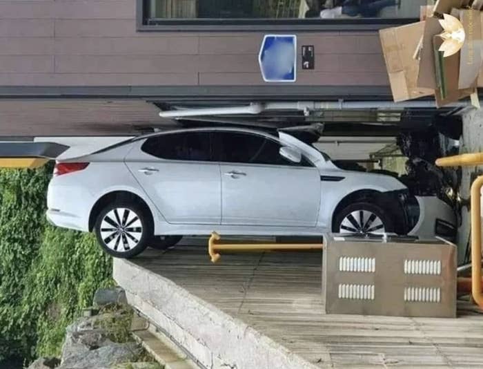 A car is parked on a steep ramp extending to an entrance, with boxes and equipment stacked nearby. The car appears to have gone slightly over the edge