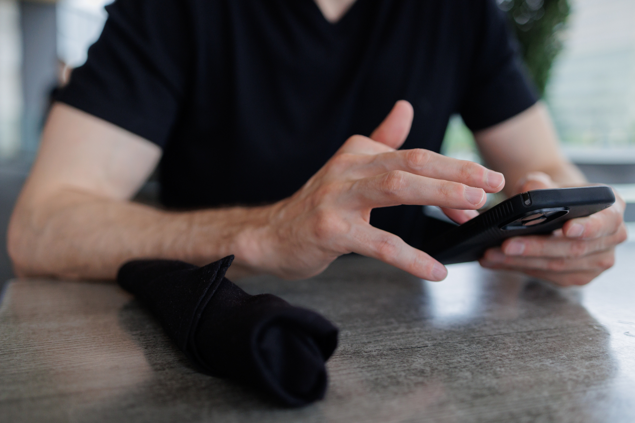 Person in a black shirt uses a smartphone at a table, with a folded cloth napkin beside them