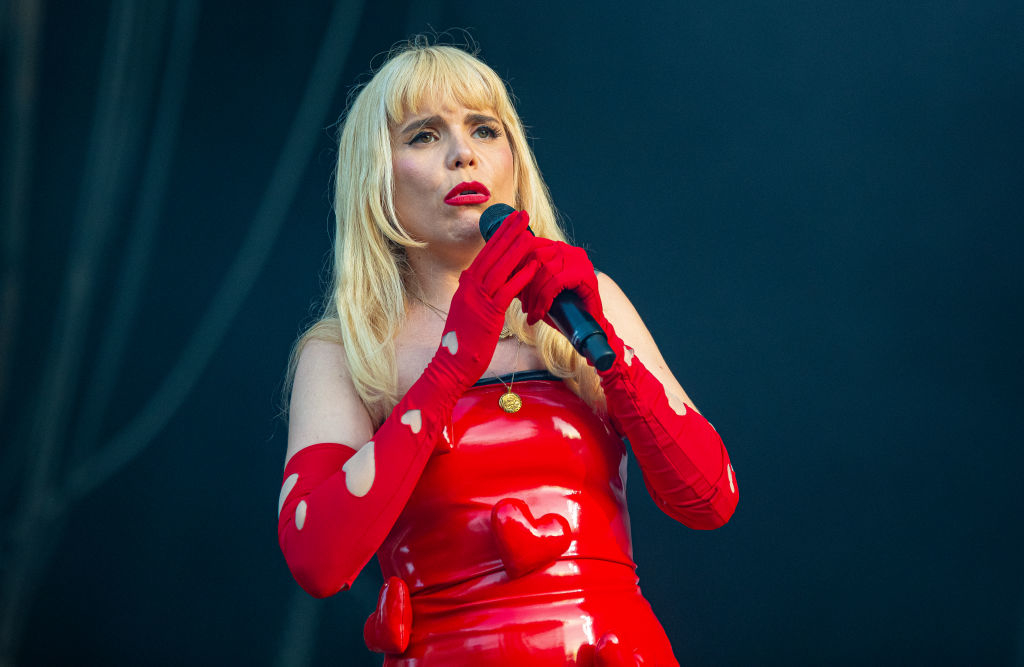 Paloma Faith performing on stage, wearing a red, form-fitting dress with matching long gloves and blonde hair