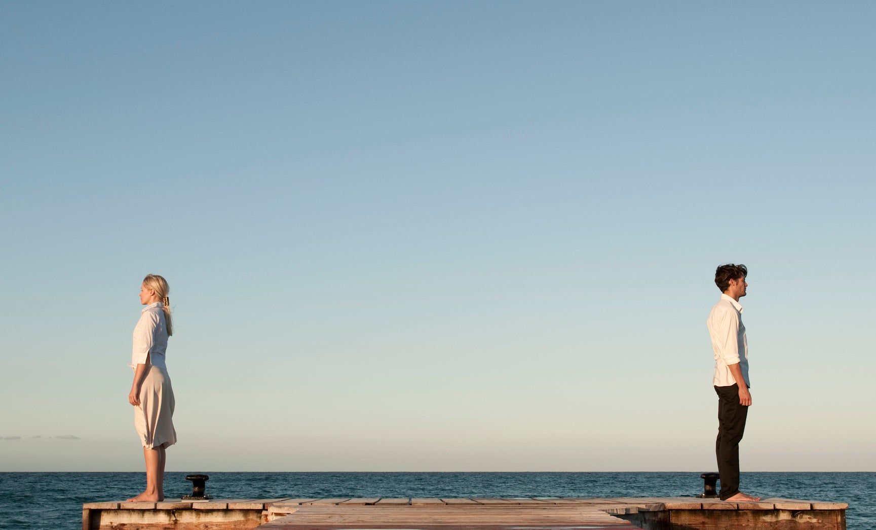 A man and woman stand on opposite ends of a dock extending into the ocean, facing away from each other, with a mountainous landscape in the background