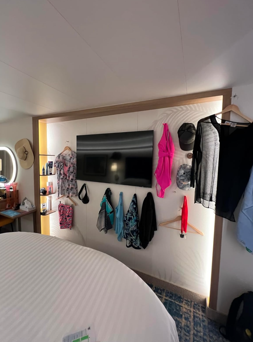 Hotel room with a TV on the wall, surrounded by various hung clothing items, including swimsuits and shirts. Bed visible in the foreground