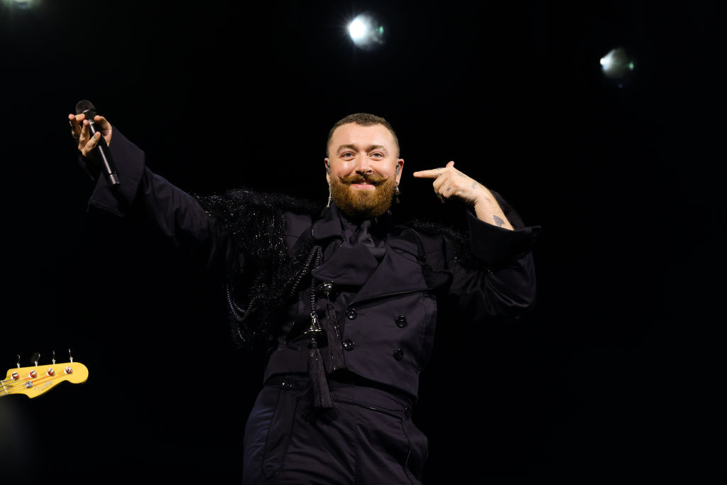 Sam Smith performing on stage, wearing a stylish black outfit and pointing to their face while holding a microphone