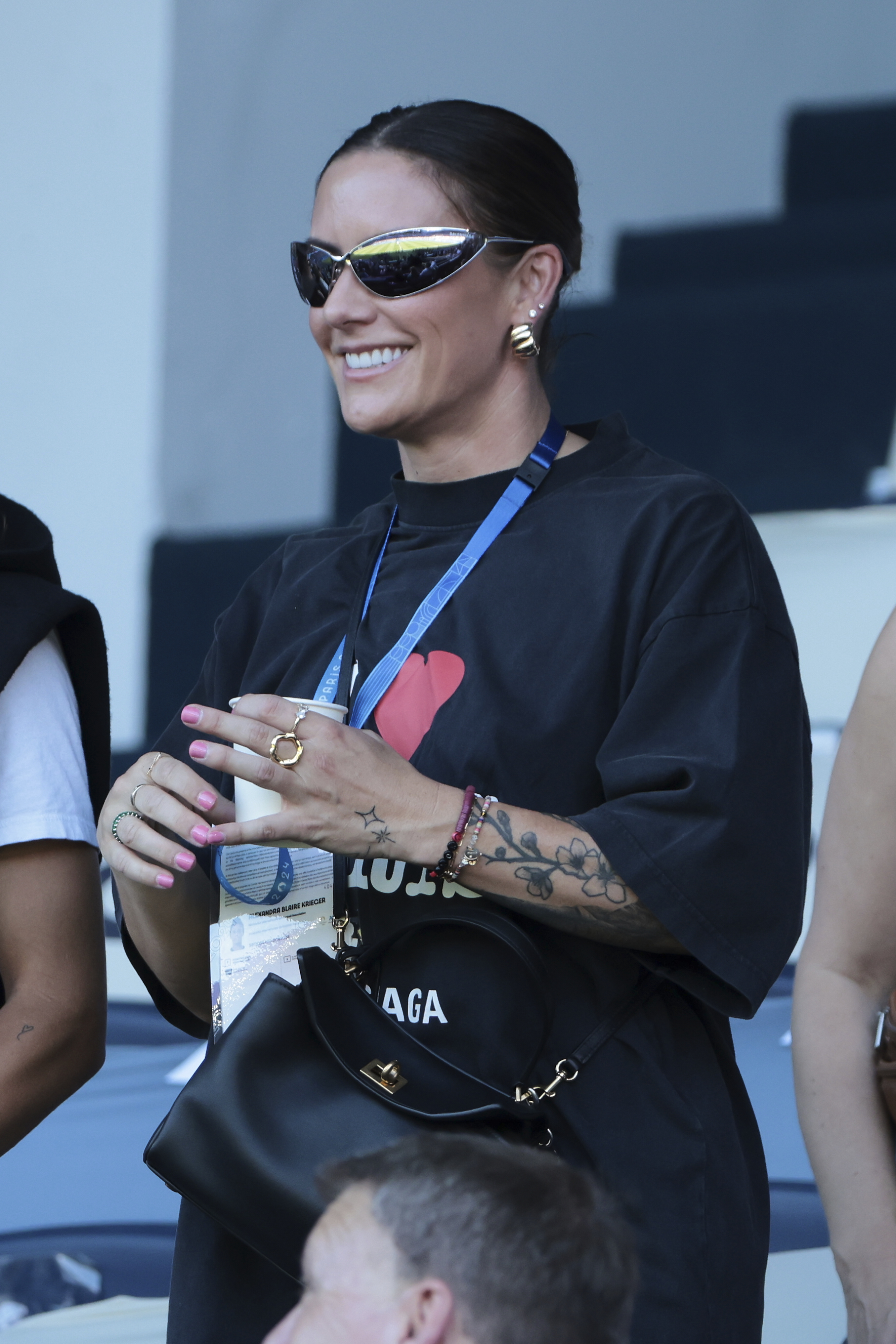 A woman smiles while wearing sunglasses, a necklace, and a black oversized shirt with tattoos visible on her right arm. She holds a Balenciaga bag