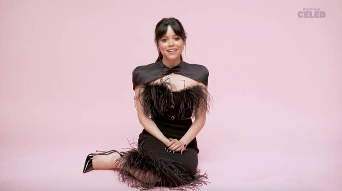 Jenna Ortega sits on the floor in a studio wearing a stylish black outfit with feather details, smiling. The background has a pink hue with &quot;BuzzFeed CELEB&quot; text
