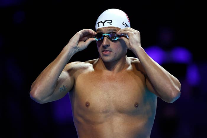 Ryan Lochte, wearing a swim cap and goggles, adjusts his goggles while standing on a swimming platform at a competitive event