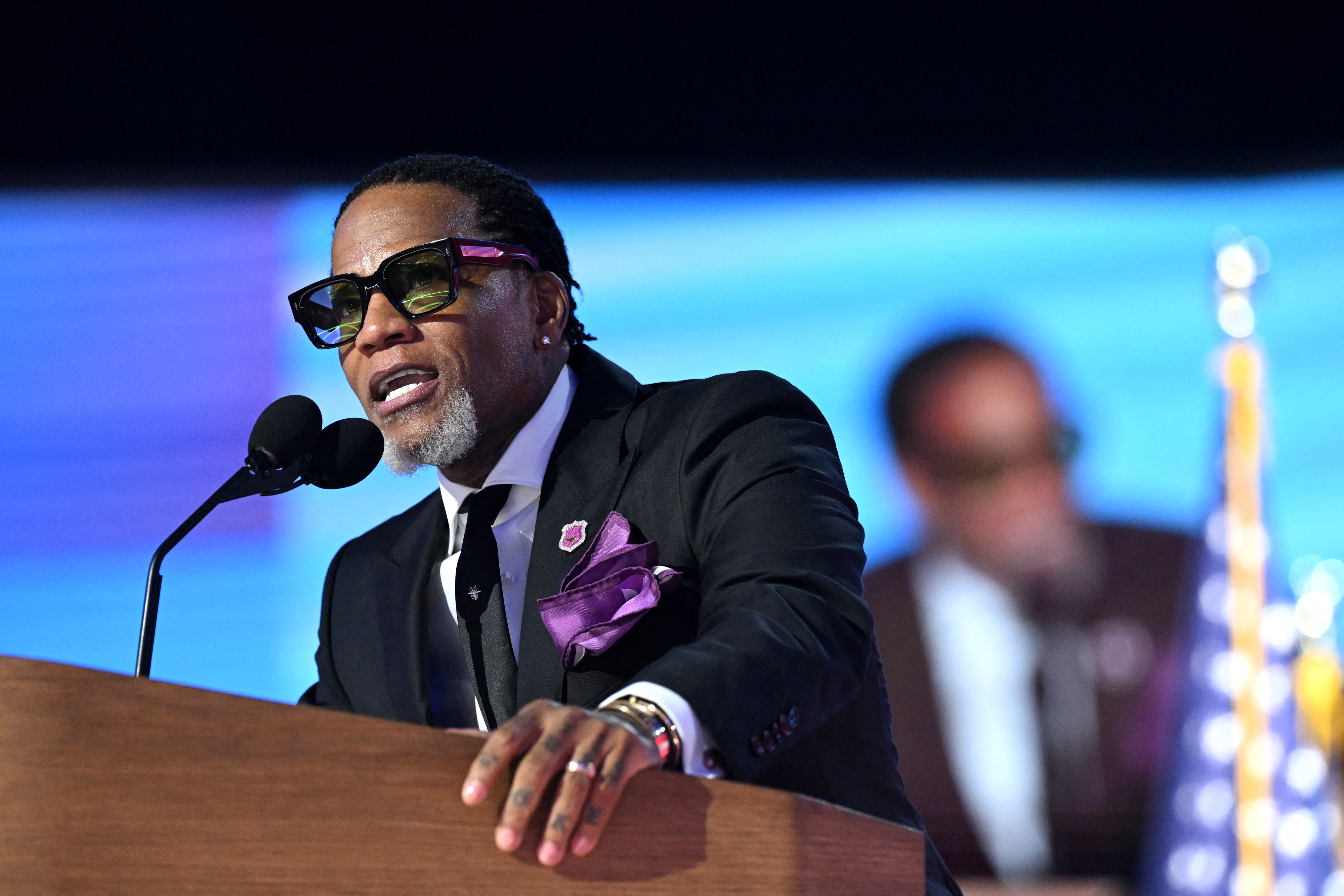 D.L. Hughley speaks at a podium wearing a dark suit and sunglasses. An unidentified man is visible in the background