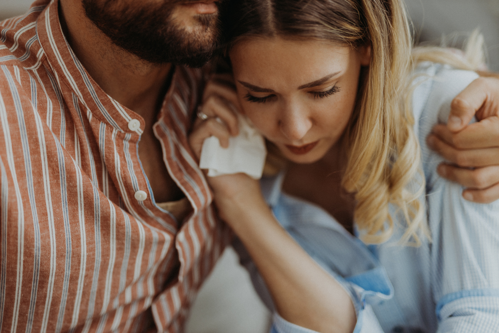 Unknown man in a striped shirt comforts a woman in a blue top who is holding a tissue and looks distressed