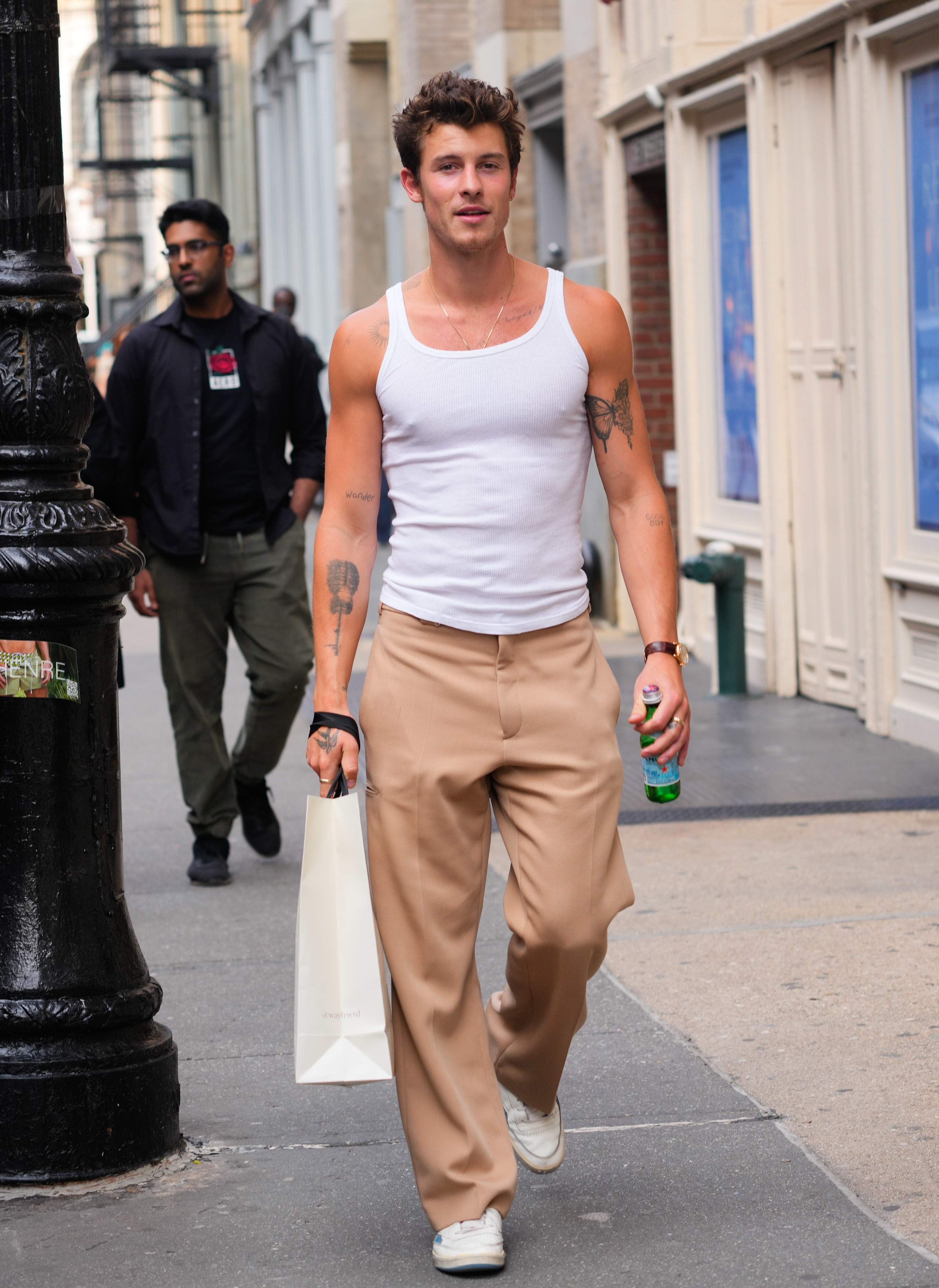 Shawn Mendes walks down a city street wearing a white tank top and beige trousers, holding a shopping bag and a drink. A person walks behind him