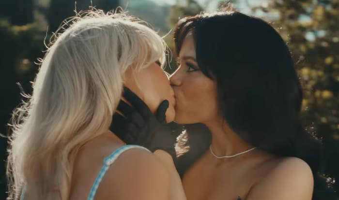 Two women sharing a kiss outdoors. One with blond hair wears a light top, and the other with dark hair wears a strapless outfit and necklace. Their names are unknown