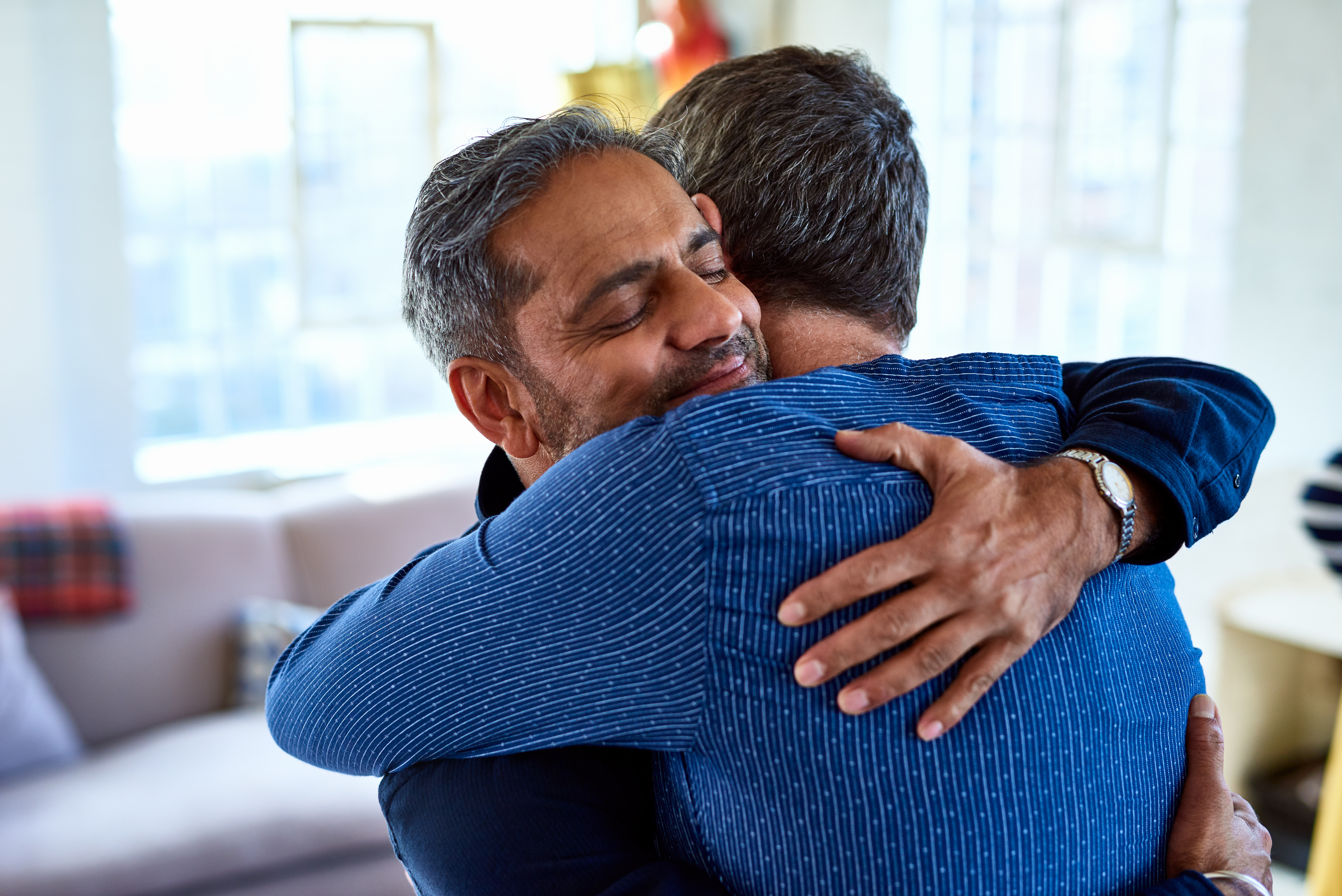 Two men, one with gray hair and the other with short dark hair, share a warm hug in a cozy living room