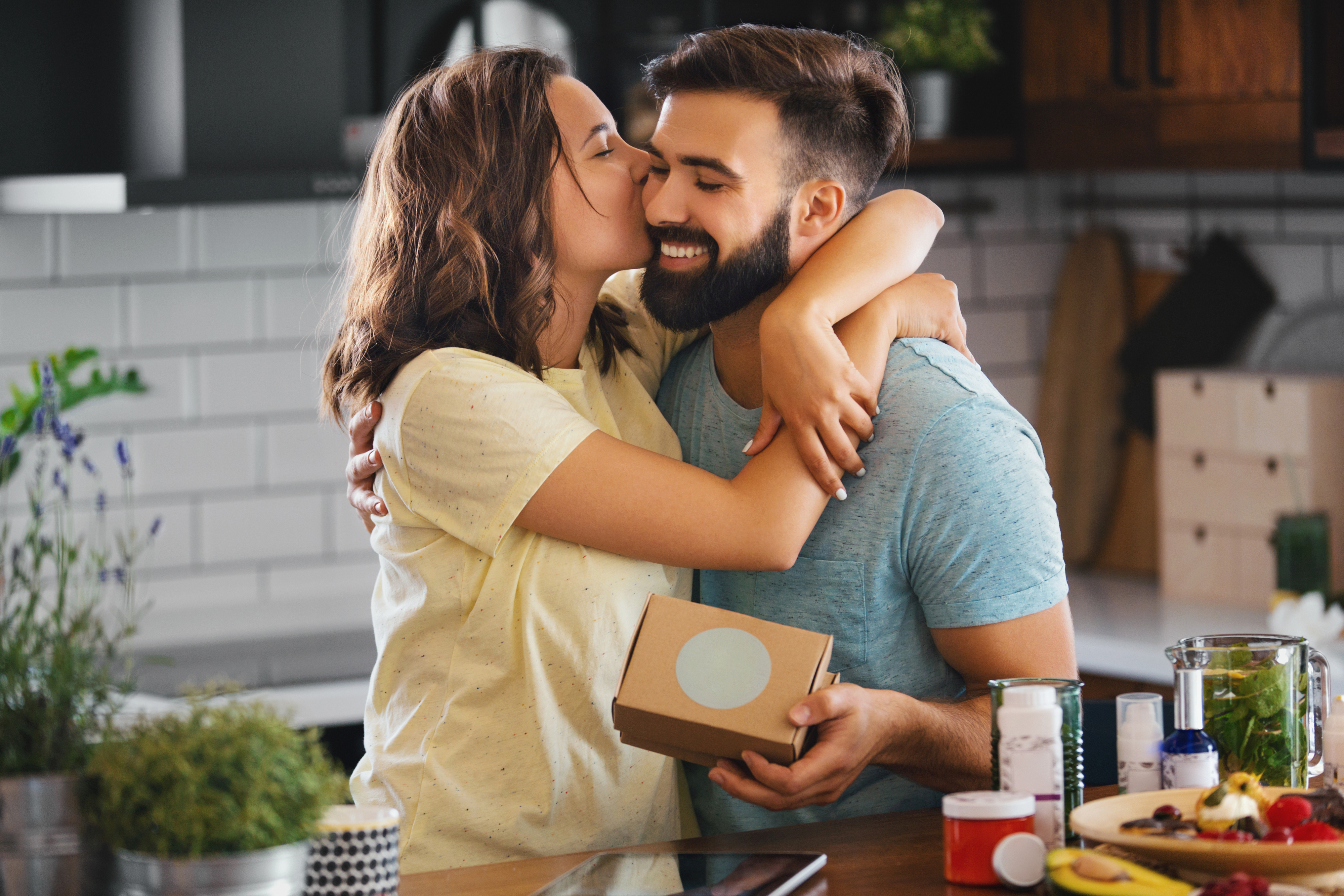 A woman kisses a smiling man on the cheek as she embraces him in a kitchen. The man holds a small cardboard box. They appear happy and affectionate