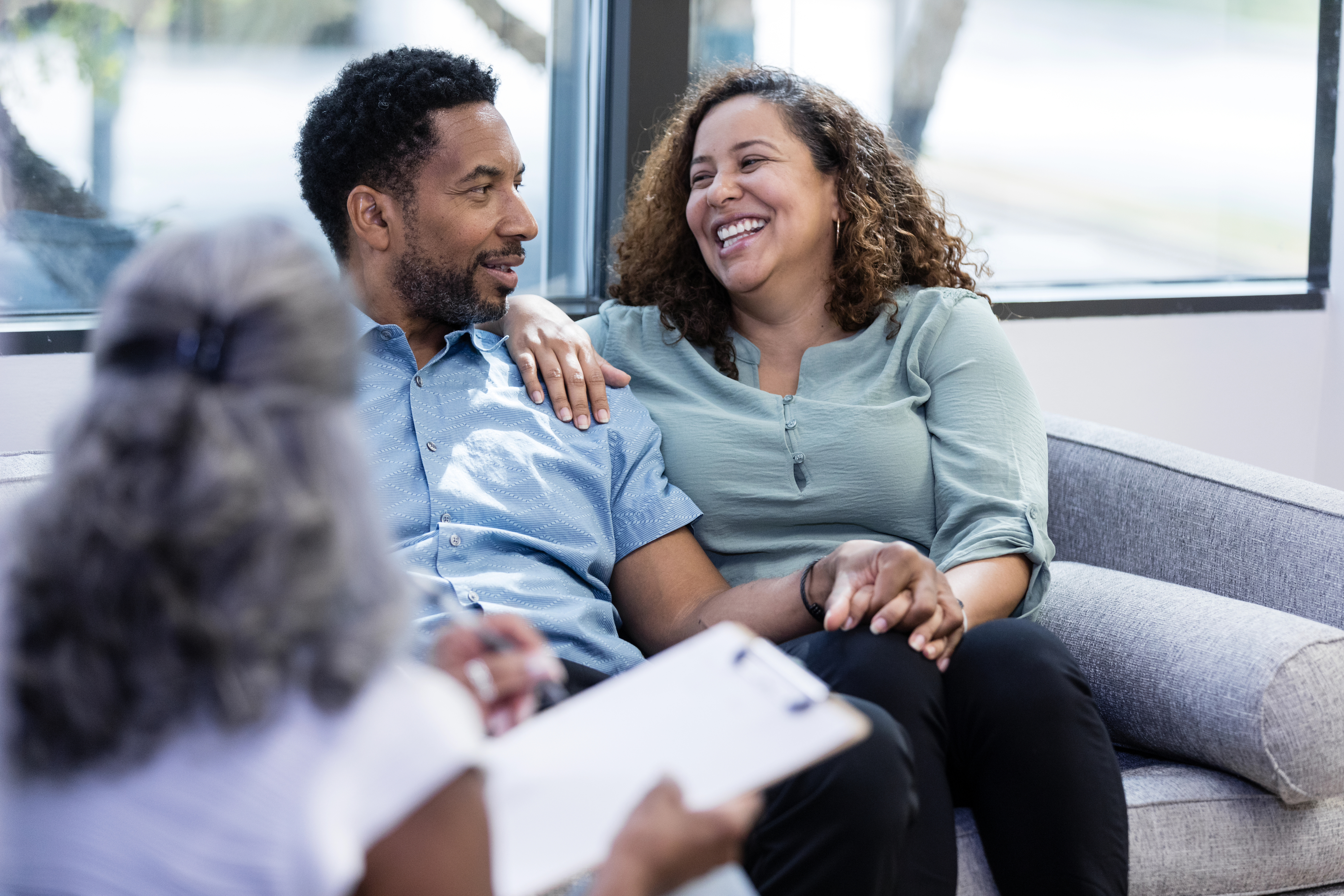 A couple sits close on a couch during a therapy session, smiling and looking at each other lovingly. A therapist with a clipboard sits in the foreground