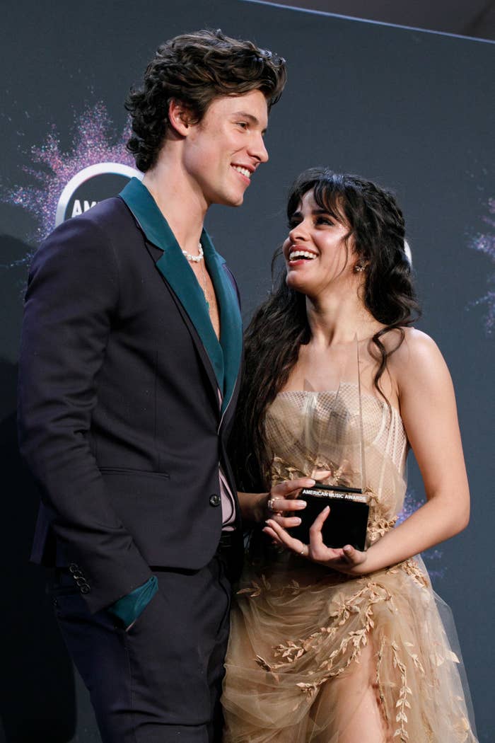 Shawn Mendes in a dark suit and Camila Cabello in an elegant dress laugh together at an awards event while she holds a trophy