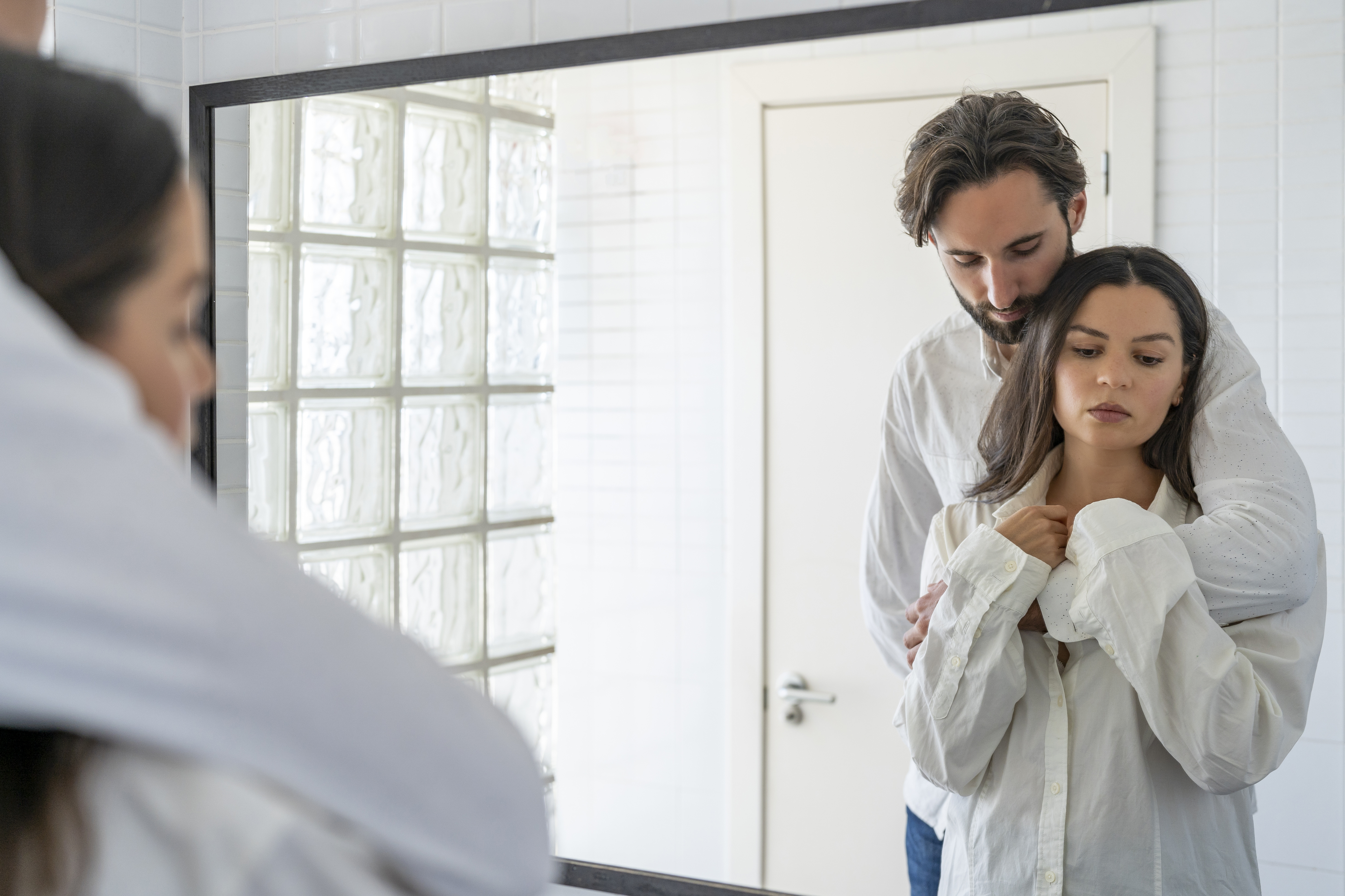 A man and a woman, both wearing white shirts, share an intimate moment in front of a mirror. The man stands behind her, gently holding her shoulders