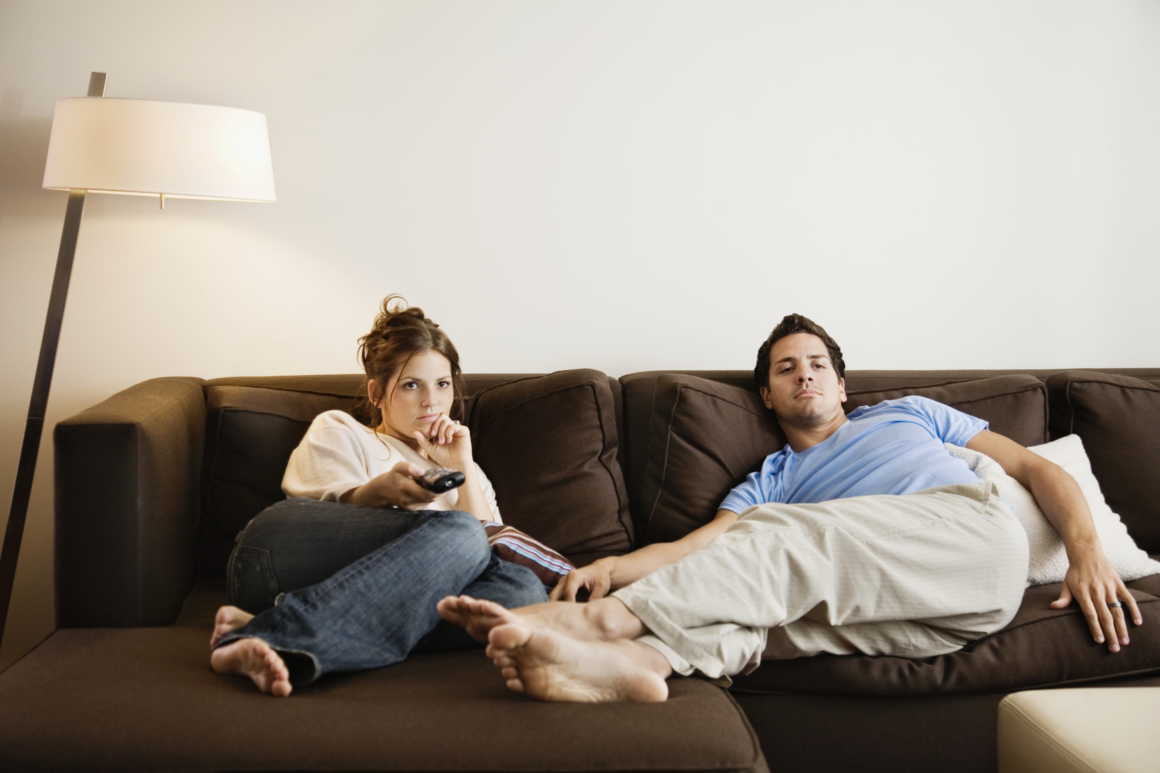 A man and woman are seated on a couch; both appear disengaged, with the woman holding a TV remote