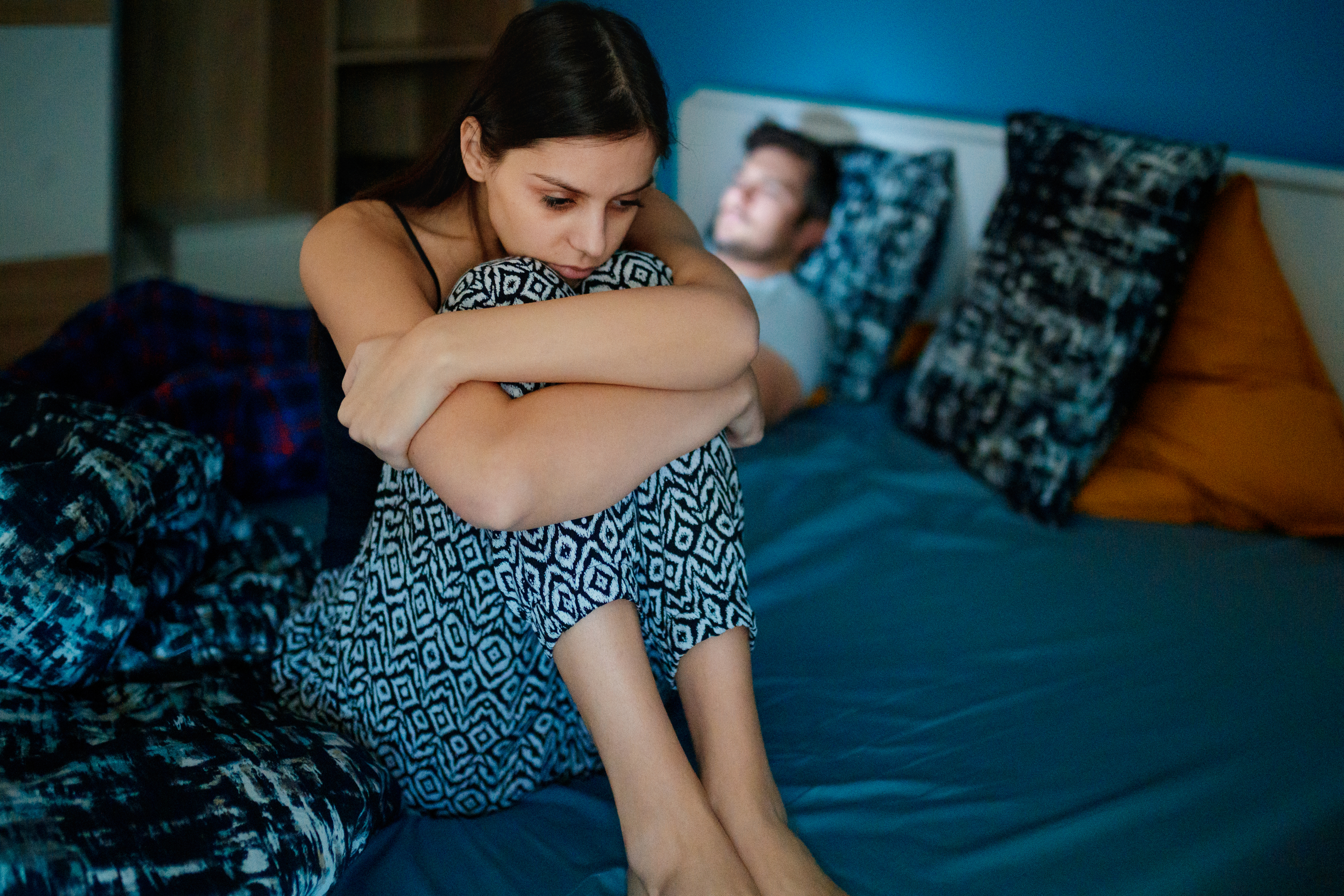 Woman sits on bed looking concerned, while a man lies in the background, seemingly asleep