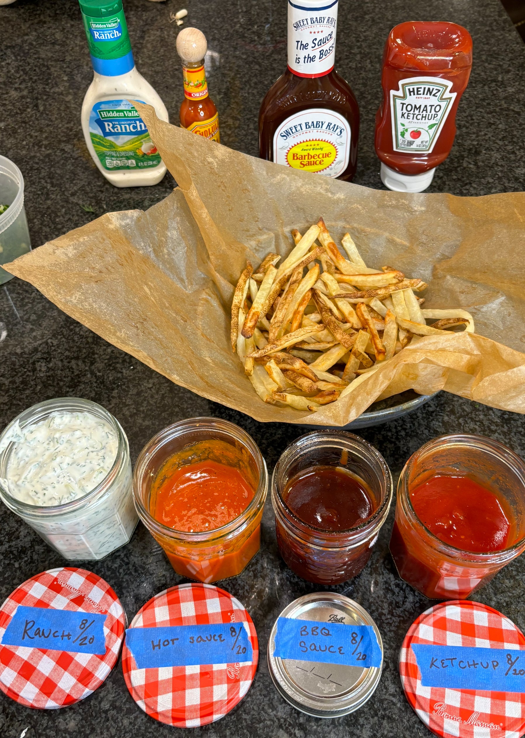 French fries on parchment paper surrounded by jars of ranch, hot sauce, BBQ sauce, and ketchup with store-bought condiments in the background
