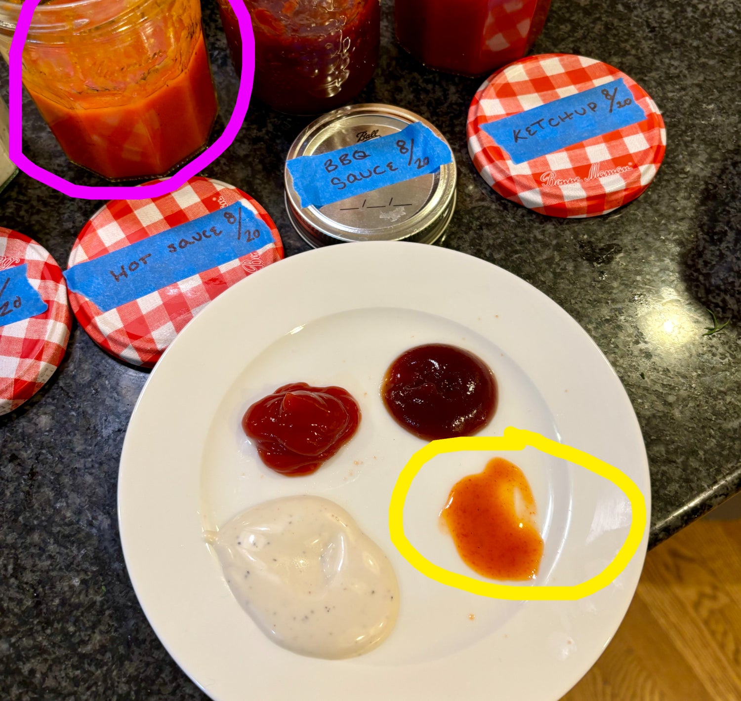 Four jars of homemade sauces with labeled lids on a counter. A white plate in the foreground shows small portions of the sauces for tasting