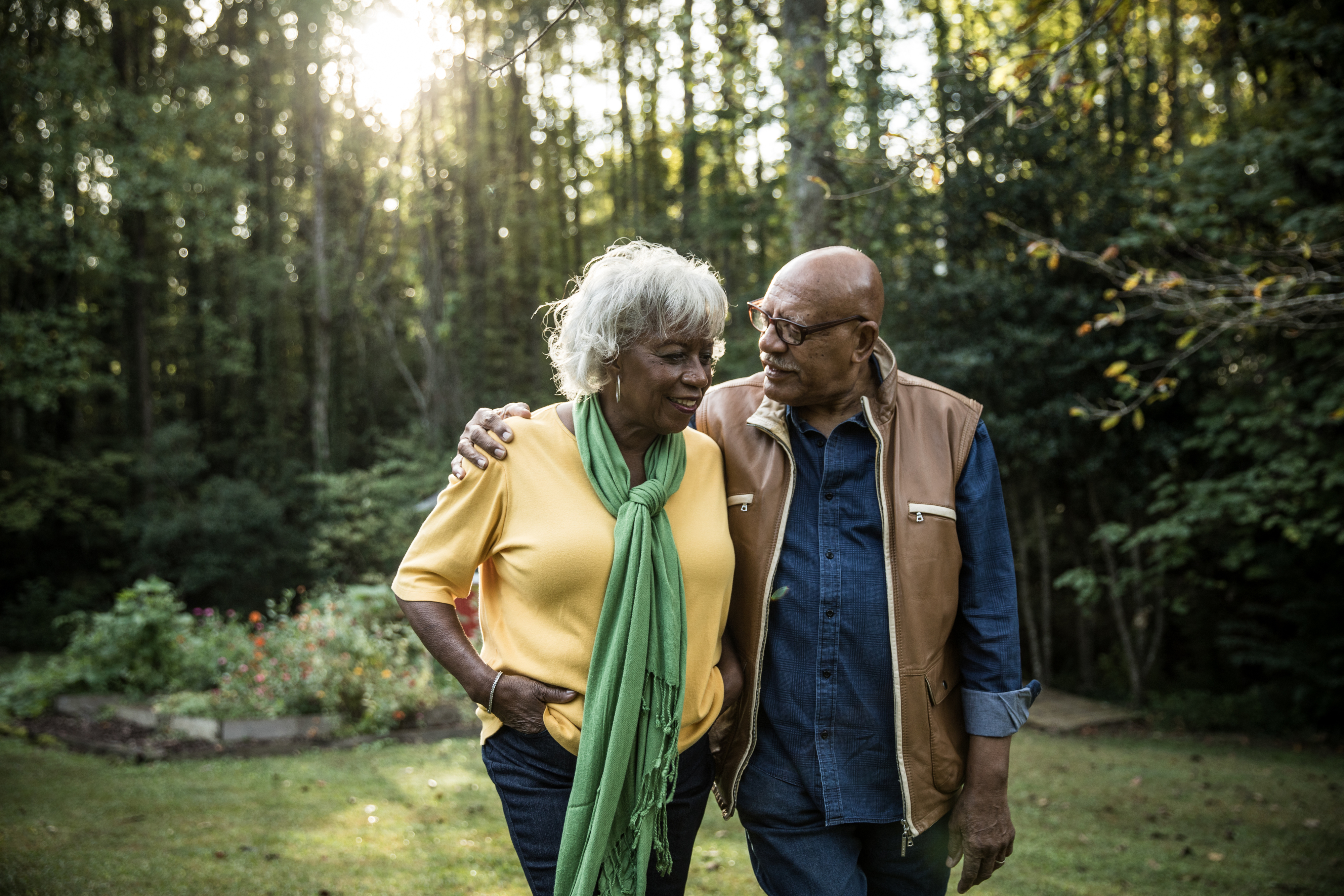 An elderly couple walk arm in arm, smiling lovingly at each other in a lush forested area