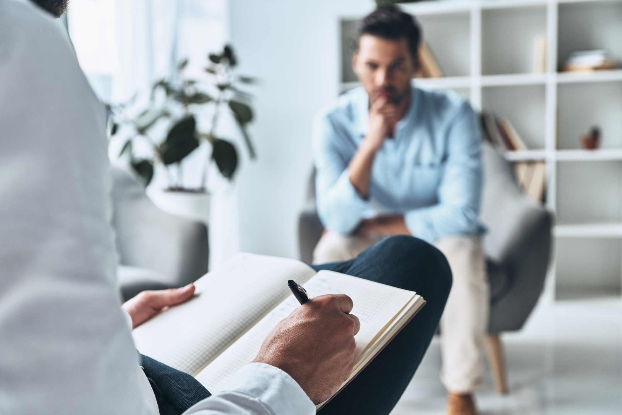 A person in casual business attire is writing in a notebook while another person sits opposite in a thoughtful pose, suggesting a counseling session