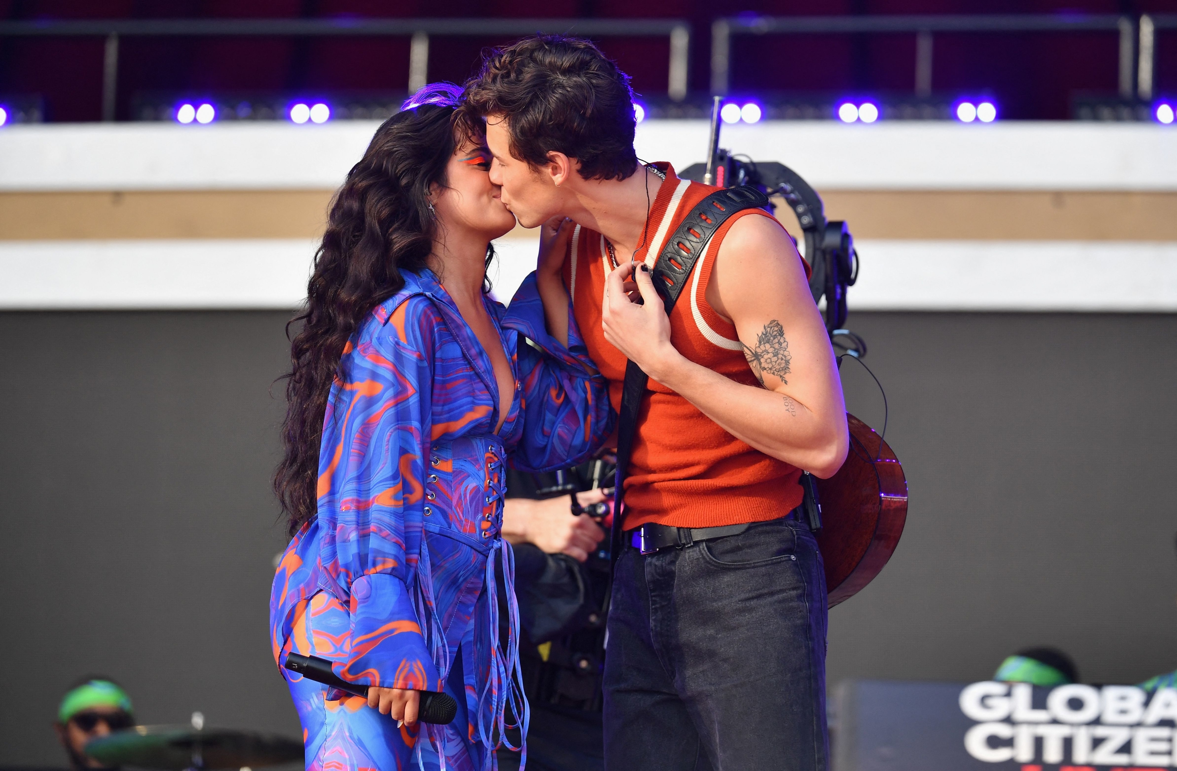 Camila Cabello in a patterned jumpsuit and Shawn Mendes in a sleeveless top kissing on stage during a performance at Global Citizen Live. An audience is in the background