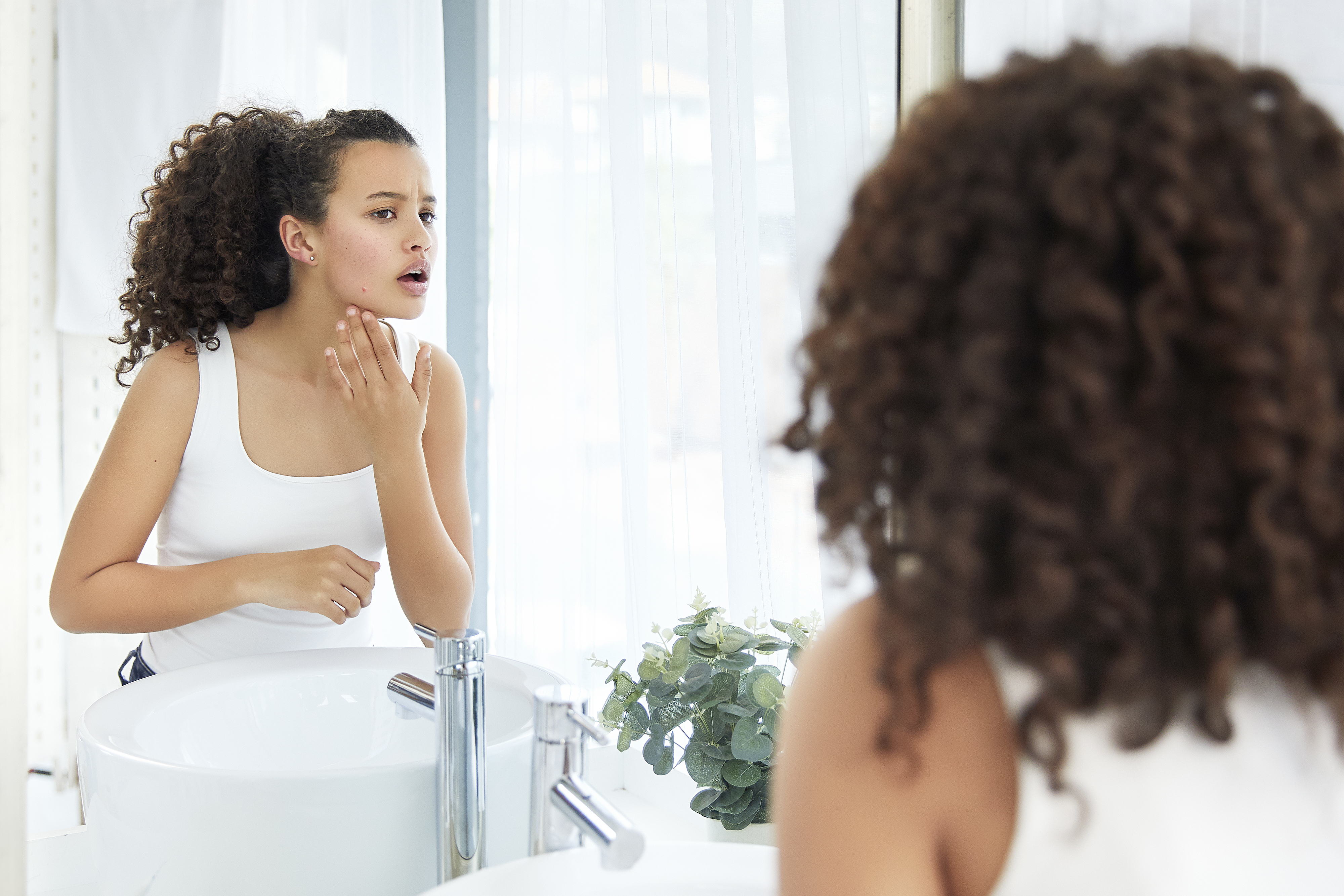 A woman with curly hair, dressed in a white tank top, examines her face in the bathroom mirror, focusing on her chin. A potted plant is visible on the sink