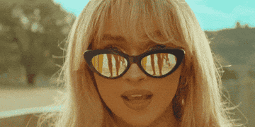 A person with long hair and sunglasses is shown up close, with reflections of people visible in the glasses. Their mouth is slightly open