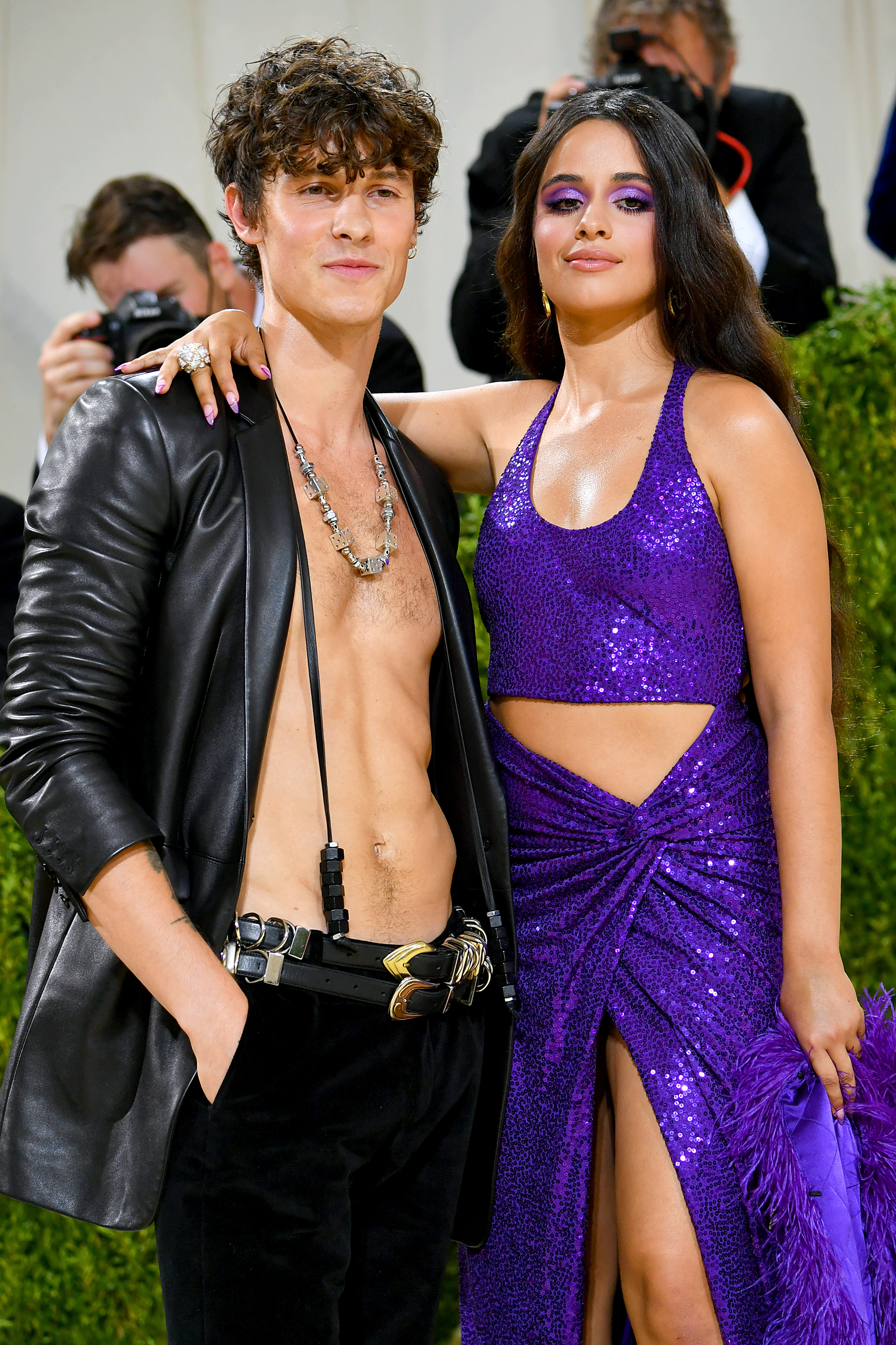 Shawn Mendes and Camila Cabello pose. Shawn wears a black leather blazer and low-rise pants, while Camila wears a purple sequined gown with a thigh-high slit