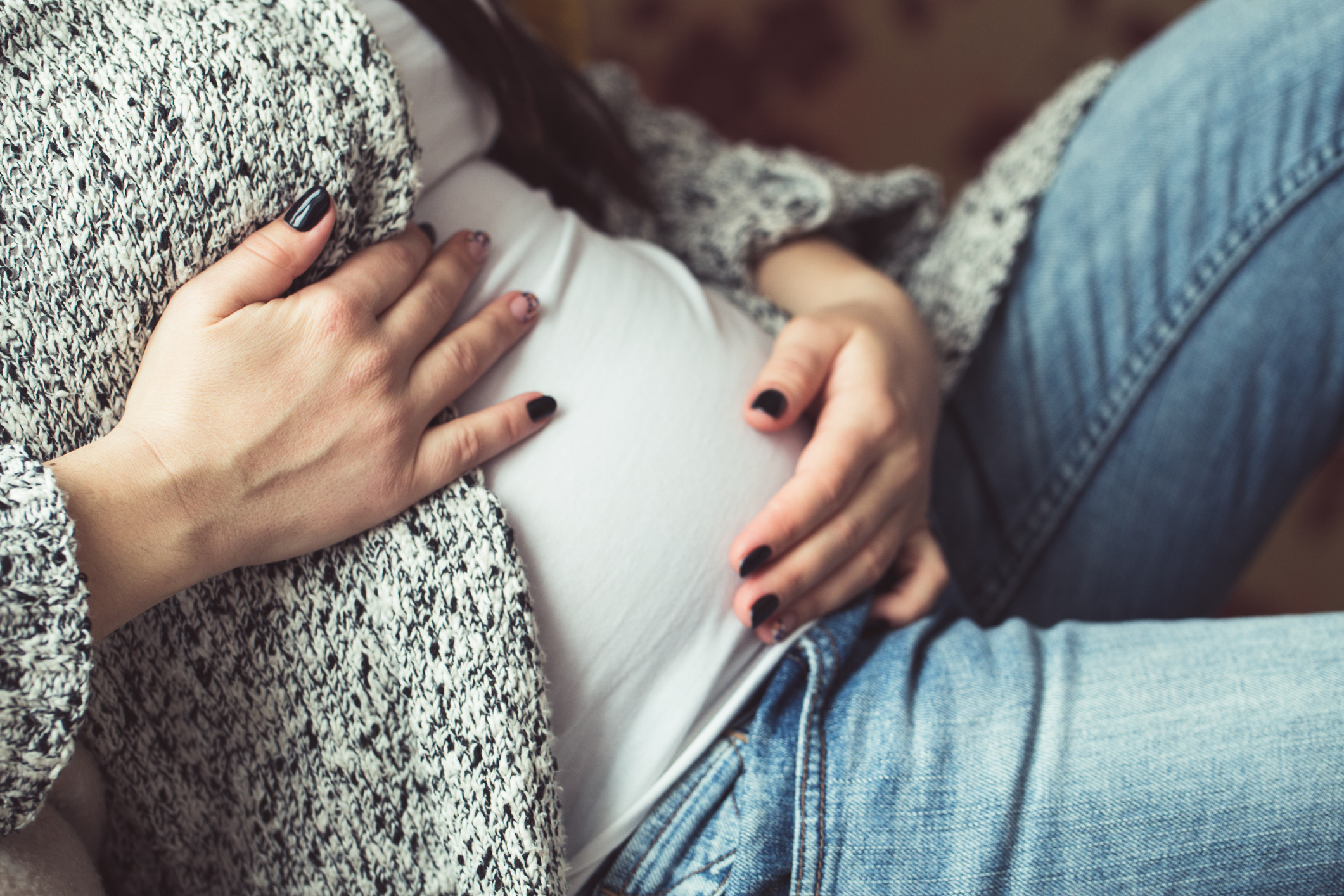 A pregnant person is sitting and gently holding their belly with both hands, wearing a sweater and jeans. The image conveys a moment of affection and anticipation