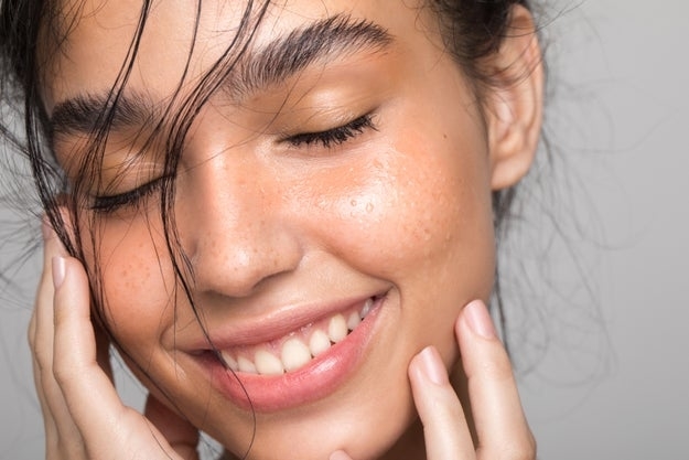 A close-up of a smiling woman with clear skin and natural makeup, gently touching her face, highlighting a beauty routine