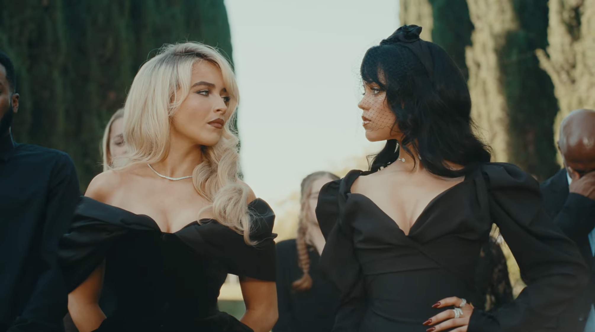 Two women, both in elegant, off-the-shoulder black dresses with puff sleeves, stand facing each other outdoors. One has long blonde hair, the other has long black hair with a veil