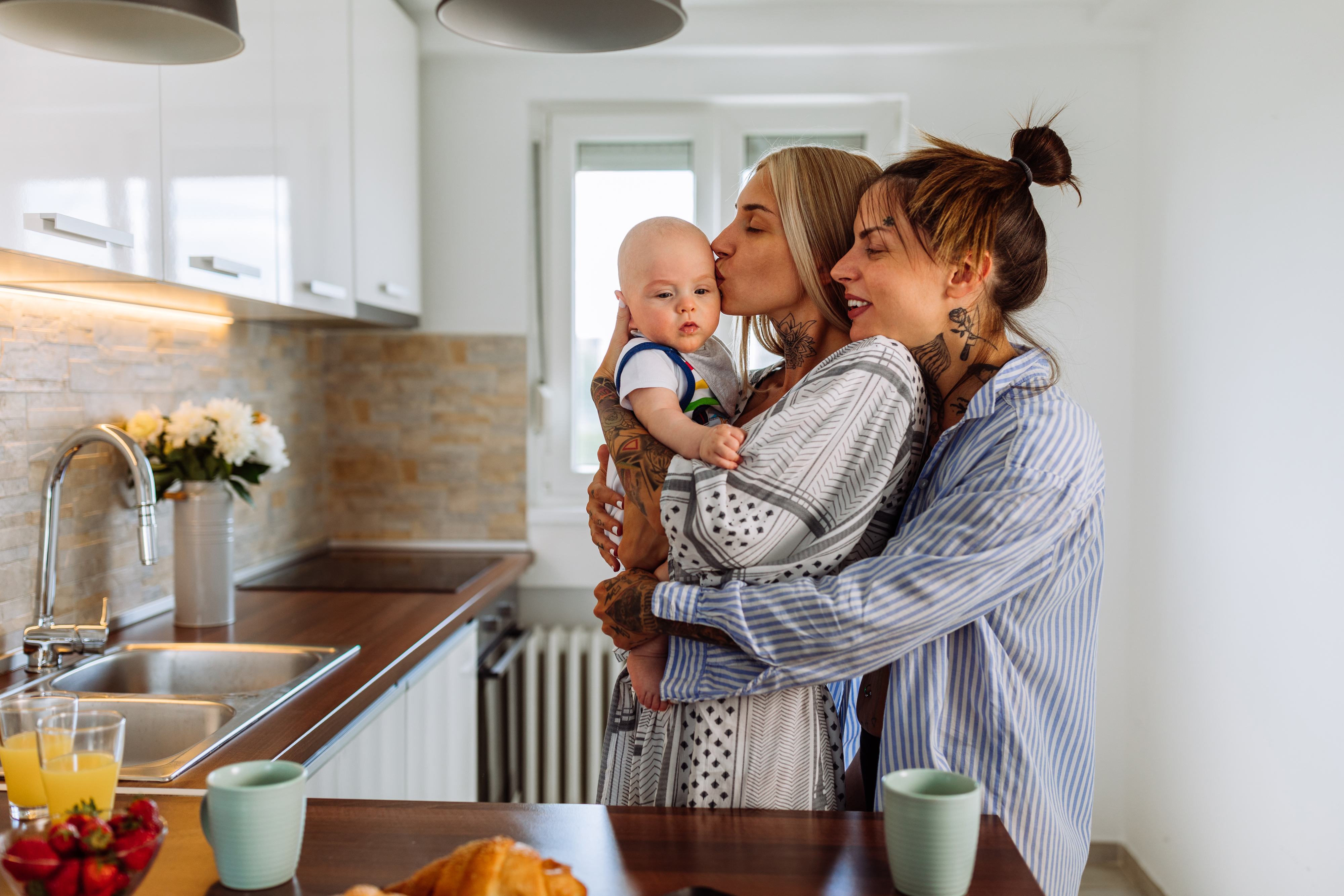 A woman with long hair kisses a baby held by another woman with tattoos. They are in a modern kitchen with a breakfast spread on the counter