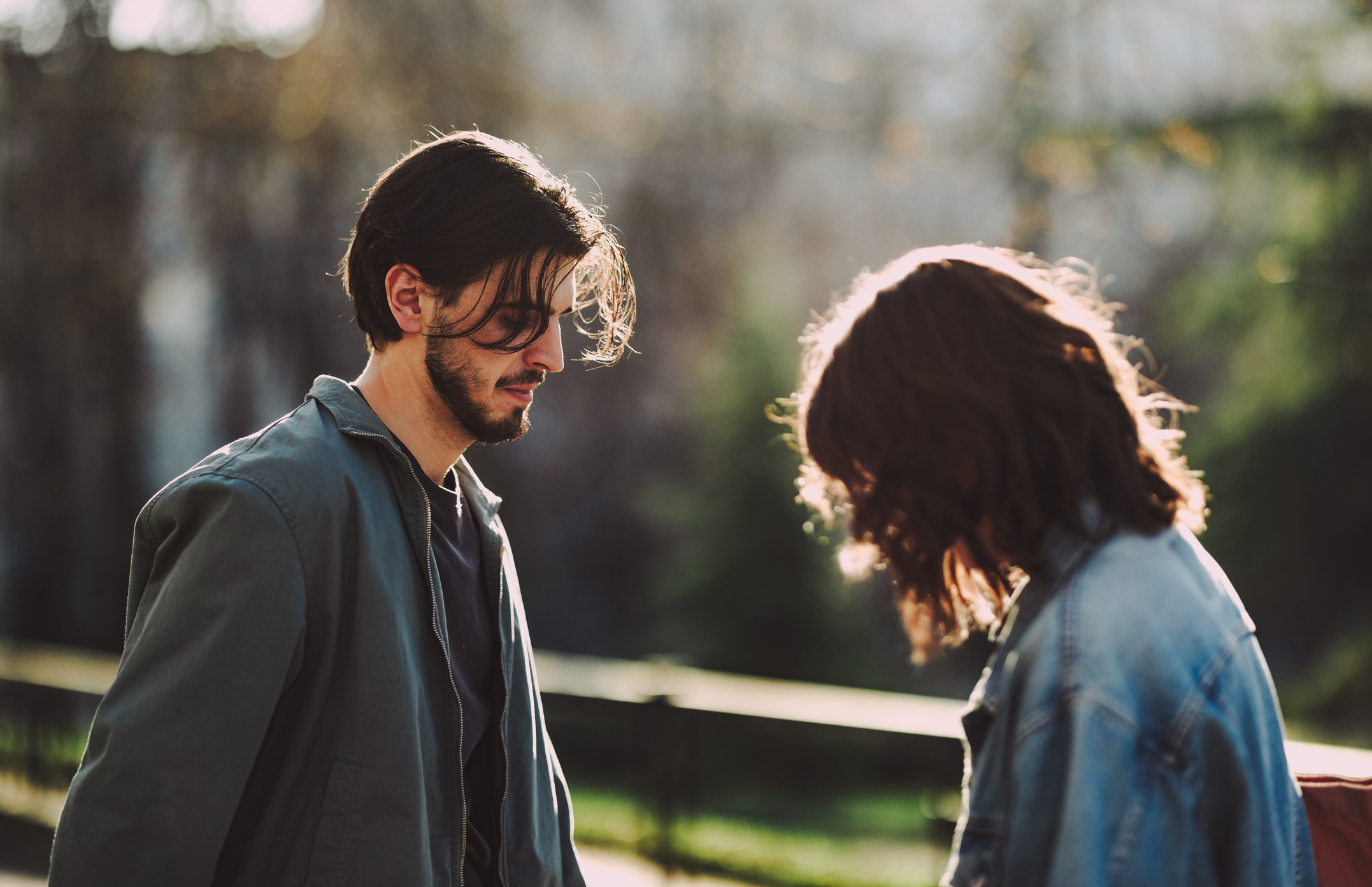 A man and woman, both wearing casual jackets, stand close to each other outdoors, their heads bent down in conversation or contemplation