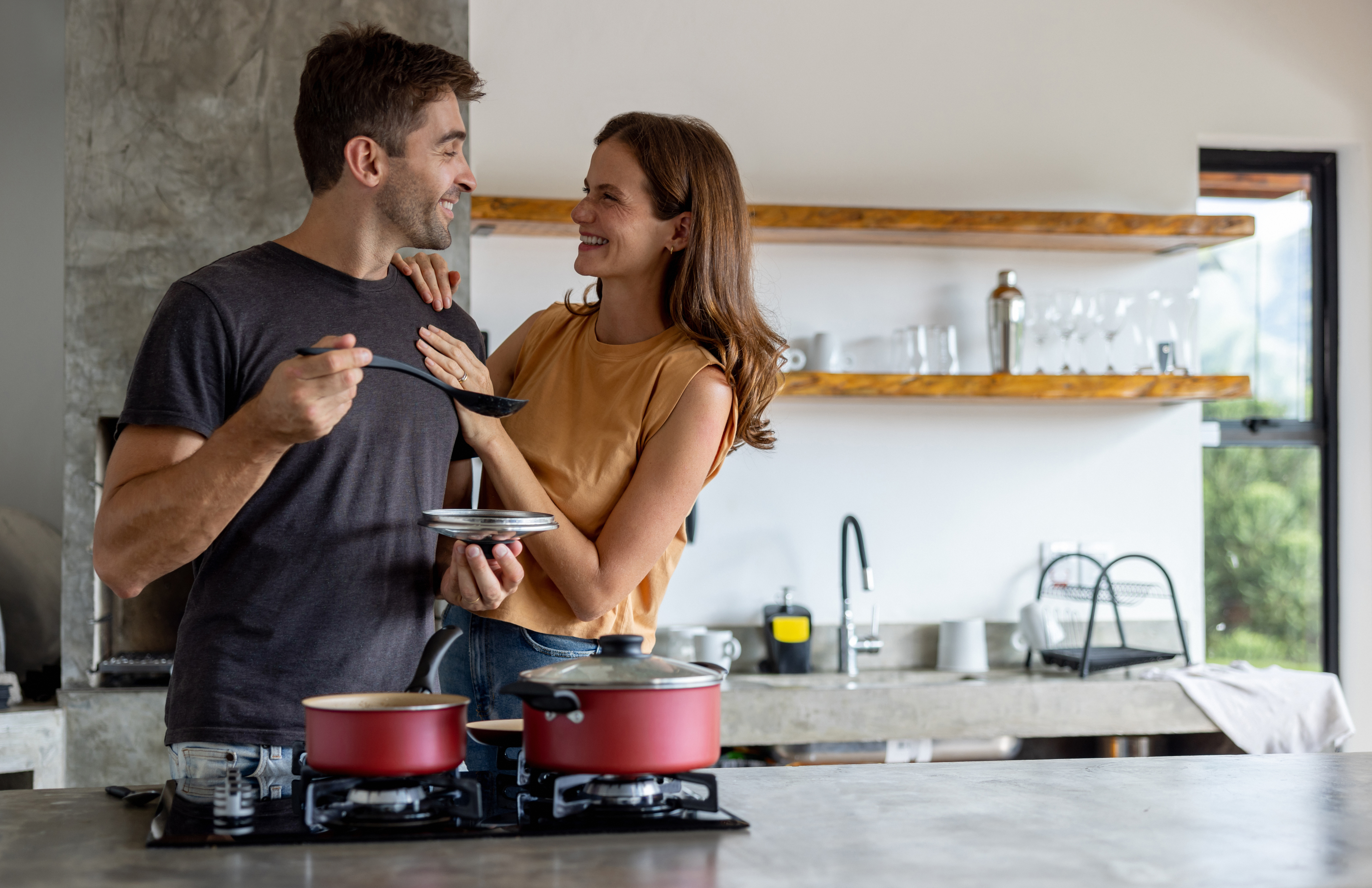 A man and a woman smile at each other while cooking together in a modern kitchen. The man holds a spoon and the woman, with her hand on her hip, looks at him lovingly