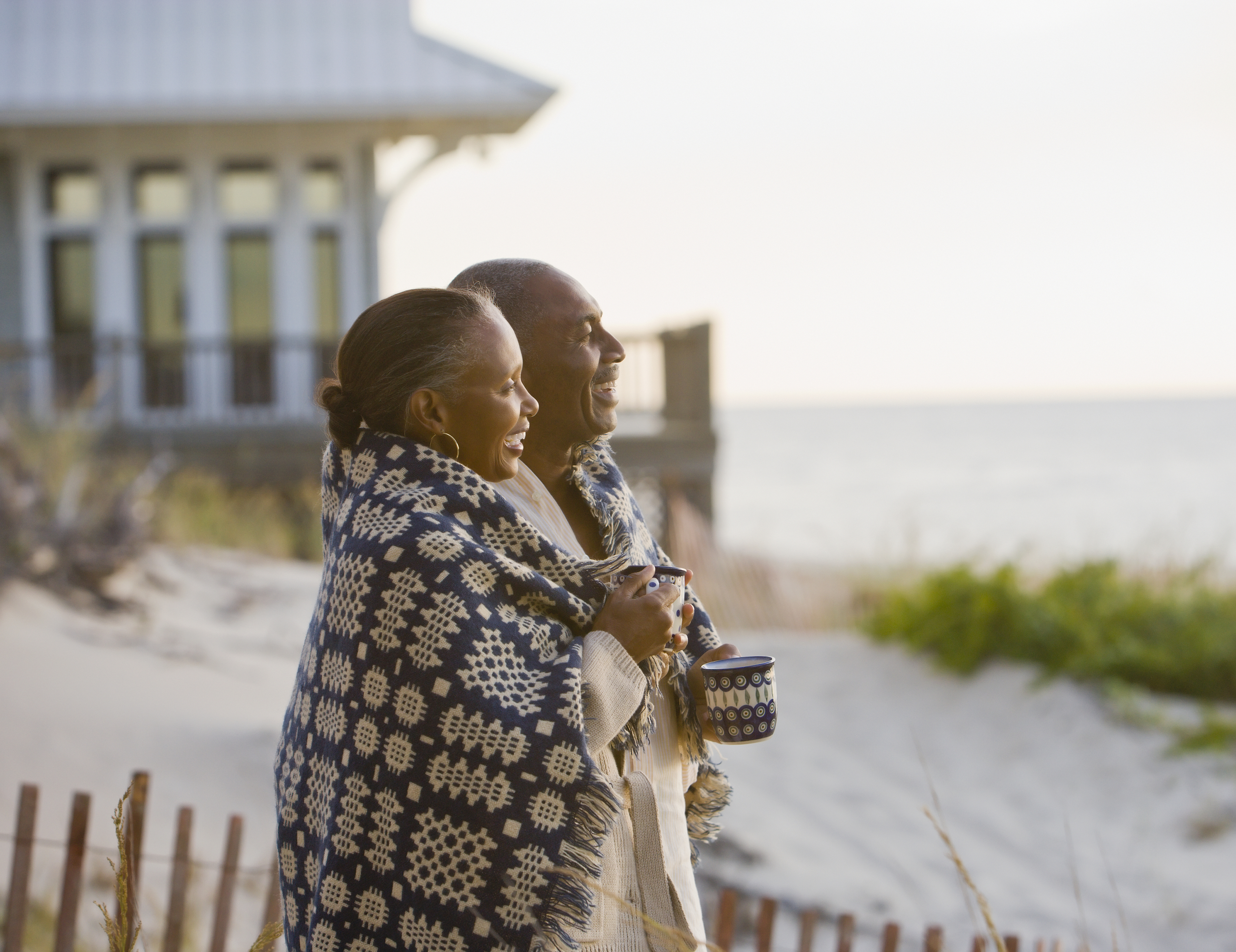 An elderly couple is standing by the beach wrapped in a patterned blanket, smiling and holding a cup, with a beachfront house in the background