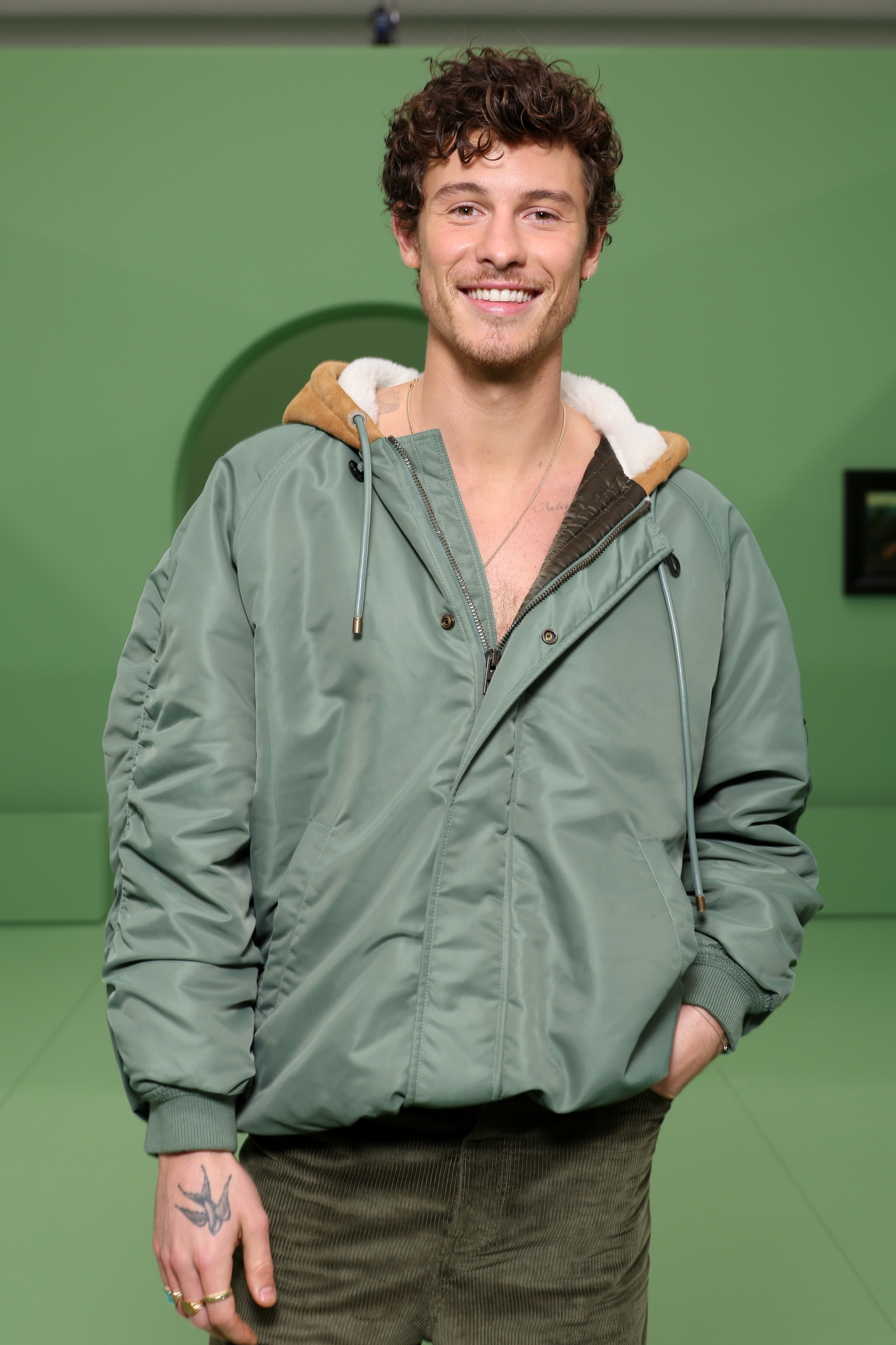 Shawn Mendes smiles while wearing a puffy zip-up jacket over a hoodie, standing with one hand in his pocket against a plain background