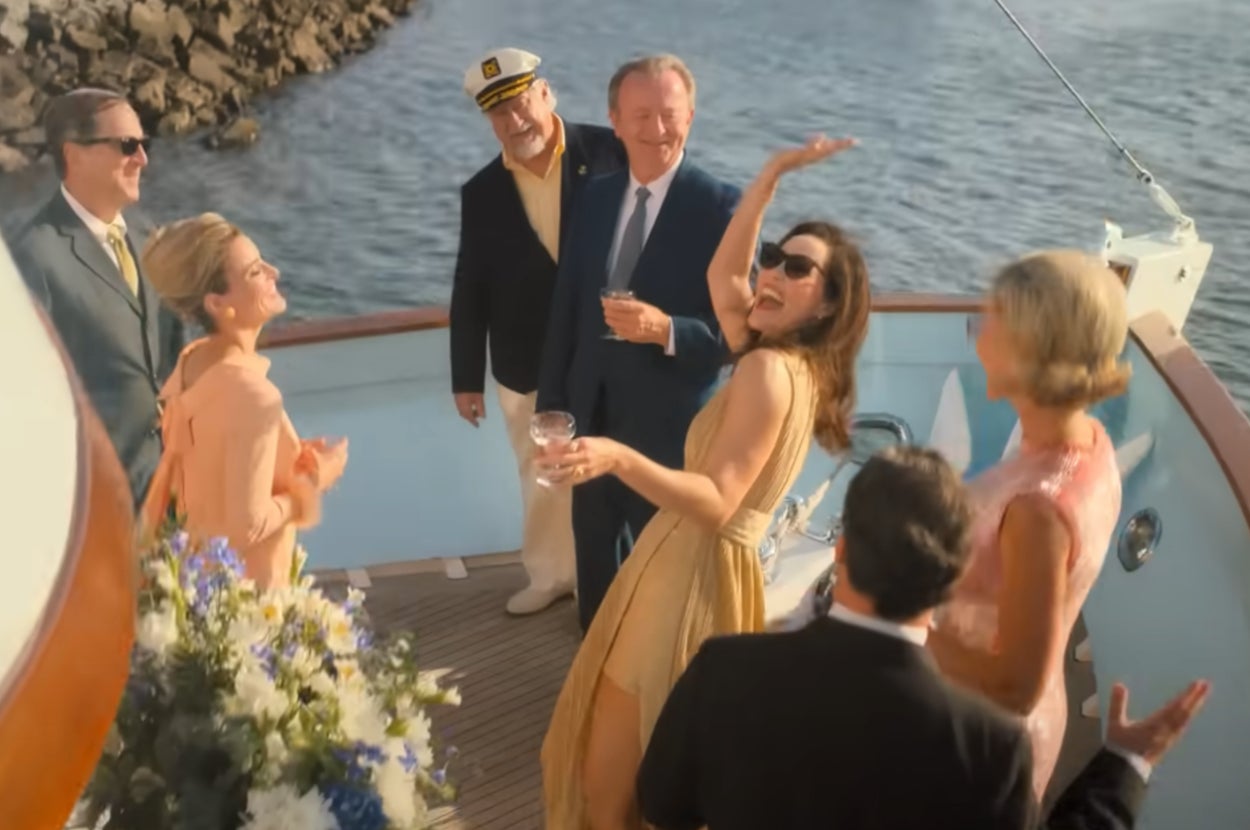 Group of people in elegant attire enjoying drinks on a yacht. Two men in captain hats, three women in dresses. Sea and dock in background.