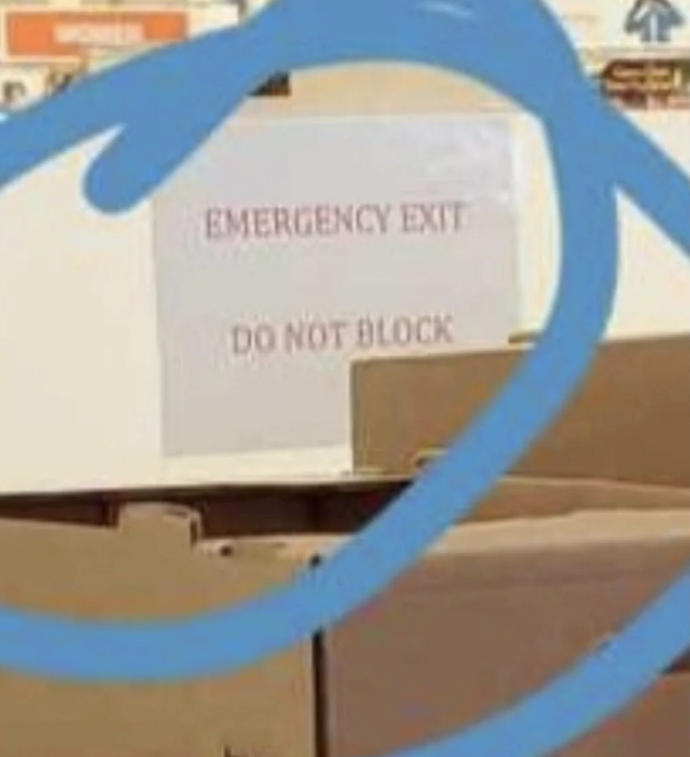 Sign reading &quot;EMERGENCY EXIT DO NOT BLOCK&quot; partially obstructed by stacked boxes in a warehouse or store