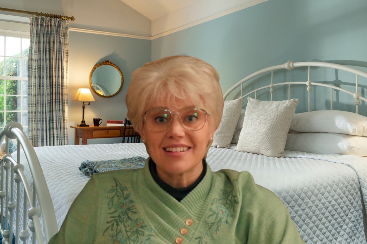 Woman wearing glasses and a green patterned sweater, smiling in front of a neatly made bed in a cozy bedroom.