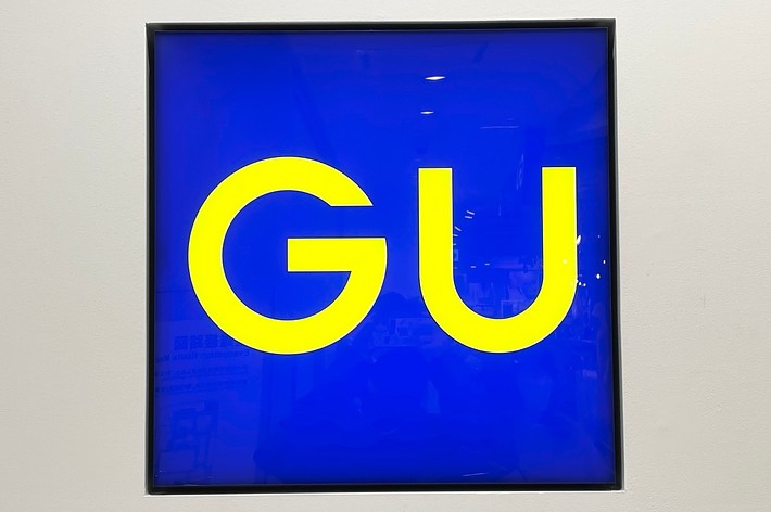 A square sign displayed on a wall shows the letters &ldquo;GU&rdquo; in bright yellow against a blue background