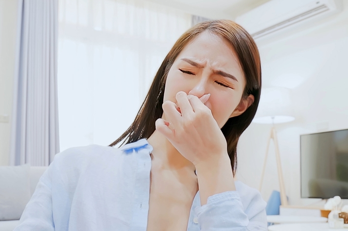 女性が室内で鼻をつまんで苦しそうな表情をしている。